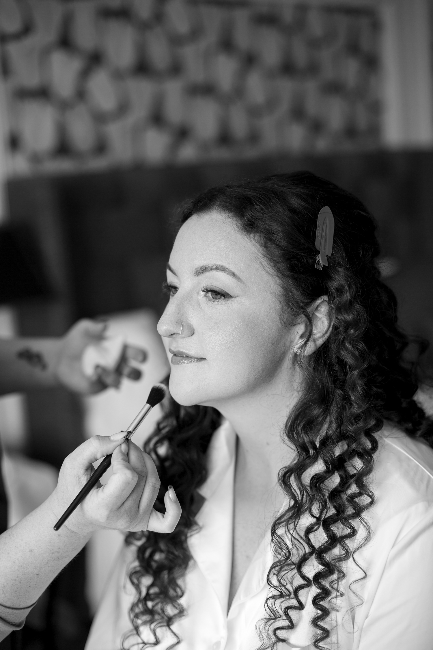 Black and white photo of a bride with curly hair getting makeup applied with a brush before her wedding.