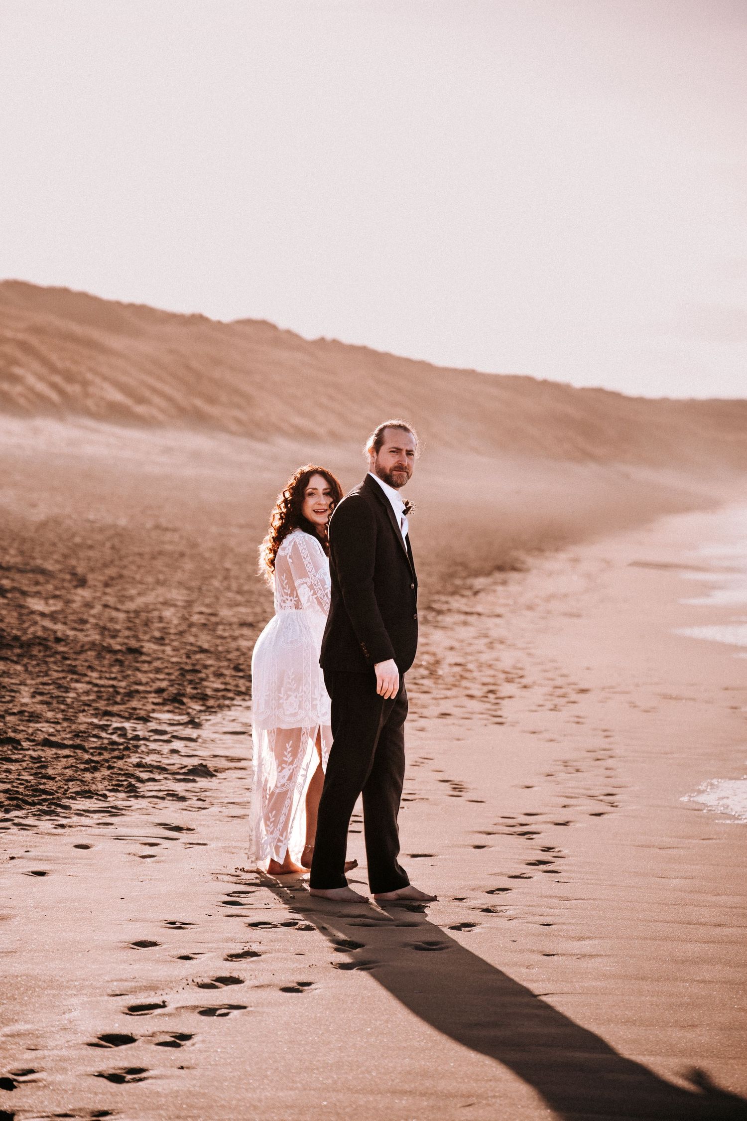 Bride and groom walking on a sandy beach at sunset, leaving footprints in the sand.