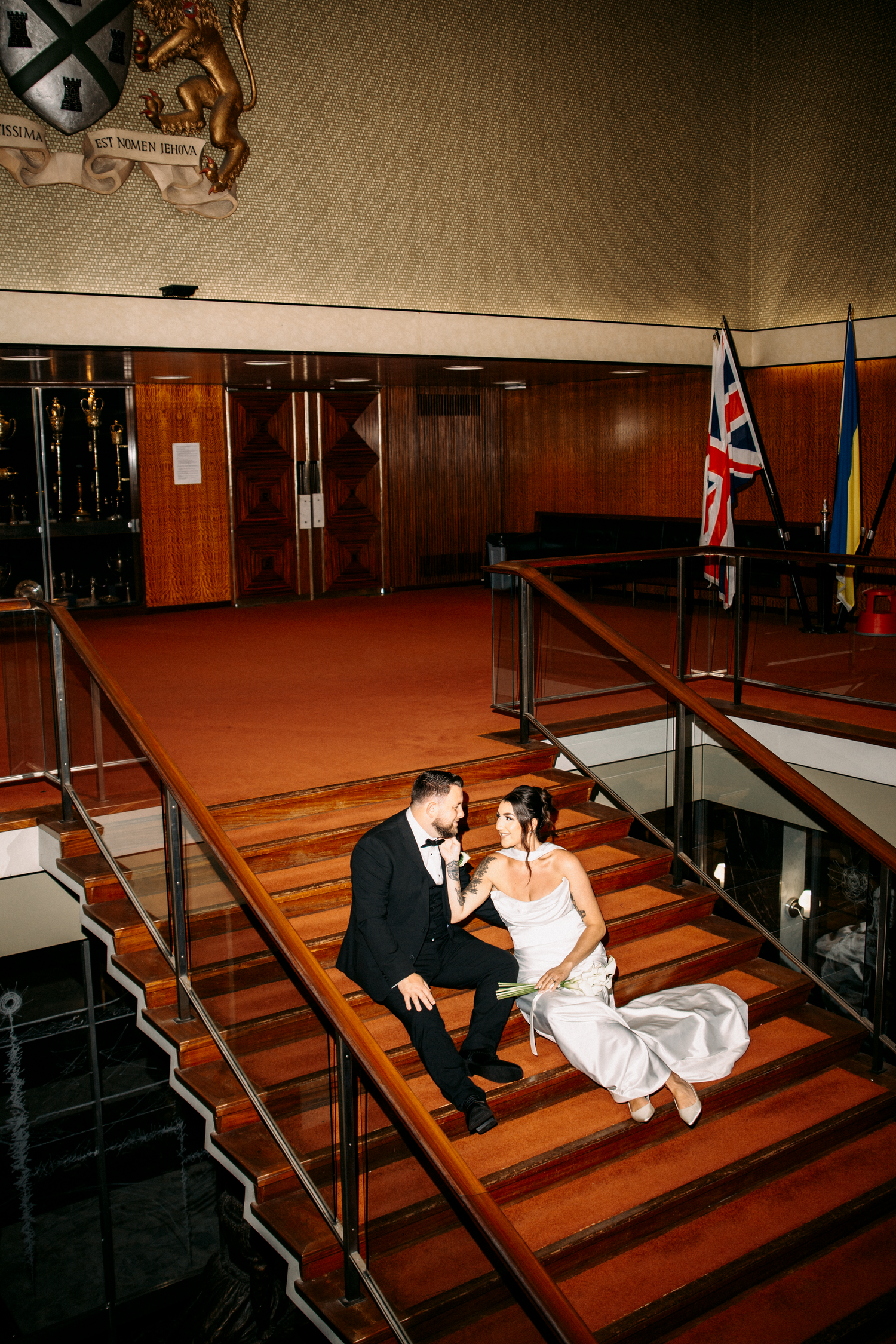 Bride and groom sit on wooden stairs in a grand building with flags and heraldic crest above.