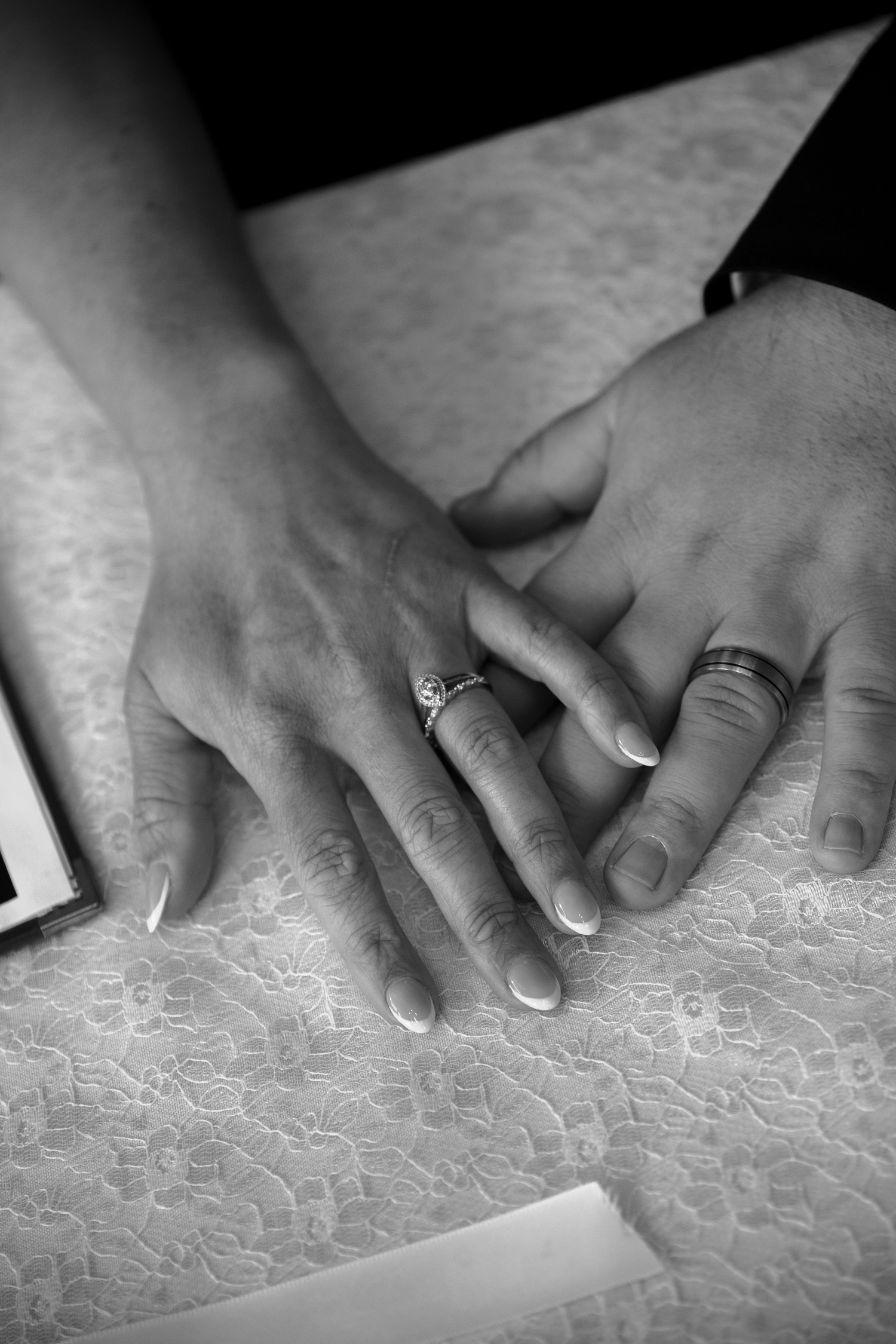 Close-up black and white photo of newlyweds hands displaying wedding rings on a lace tablecloth.