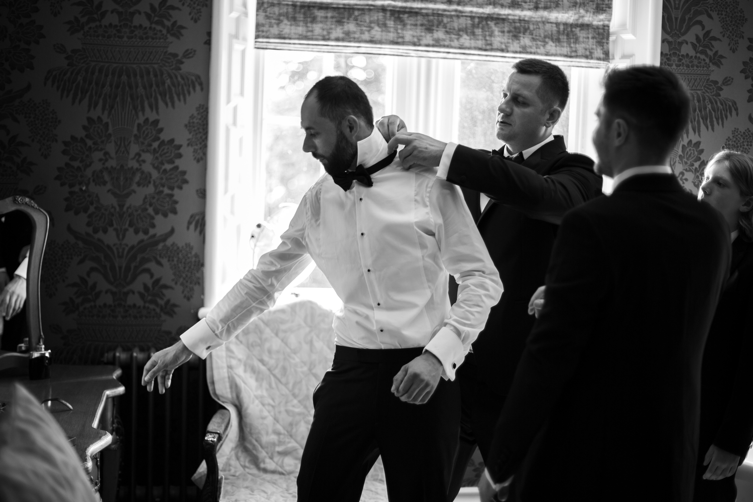 Groomsmen helping groom adjust bow tie while getting ready in elegant room on wedding day.