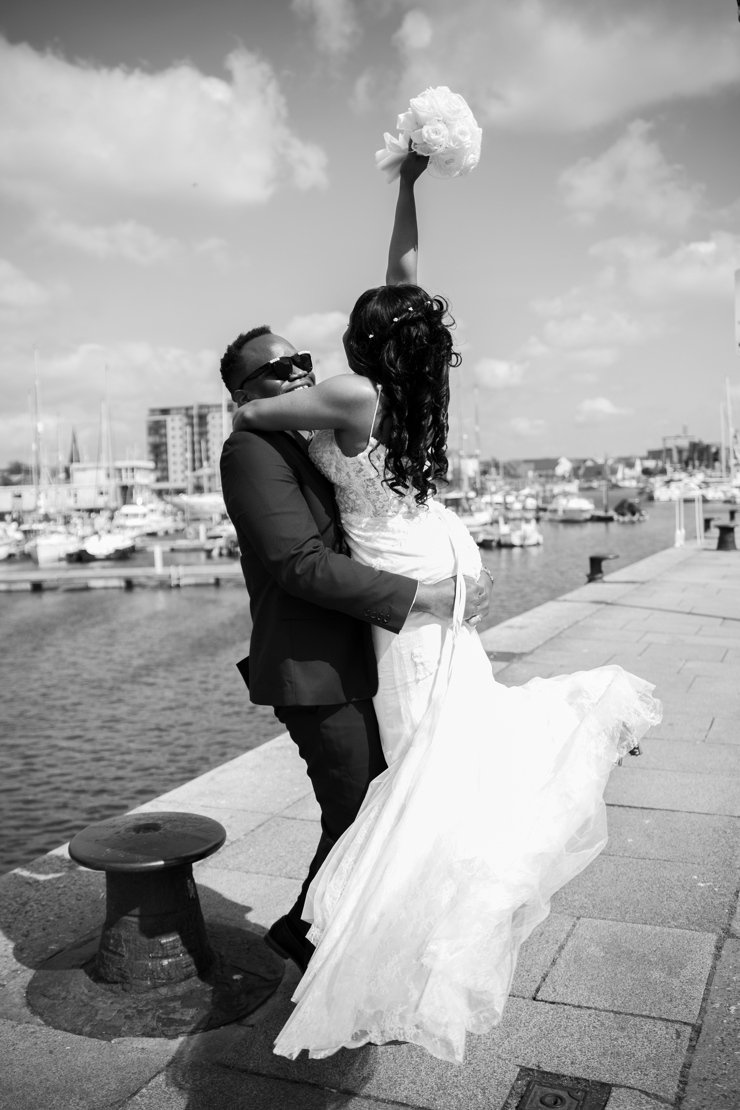 A joyful bride raises her bouquet while her groom dips her at a sunny harbor in black and white.