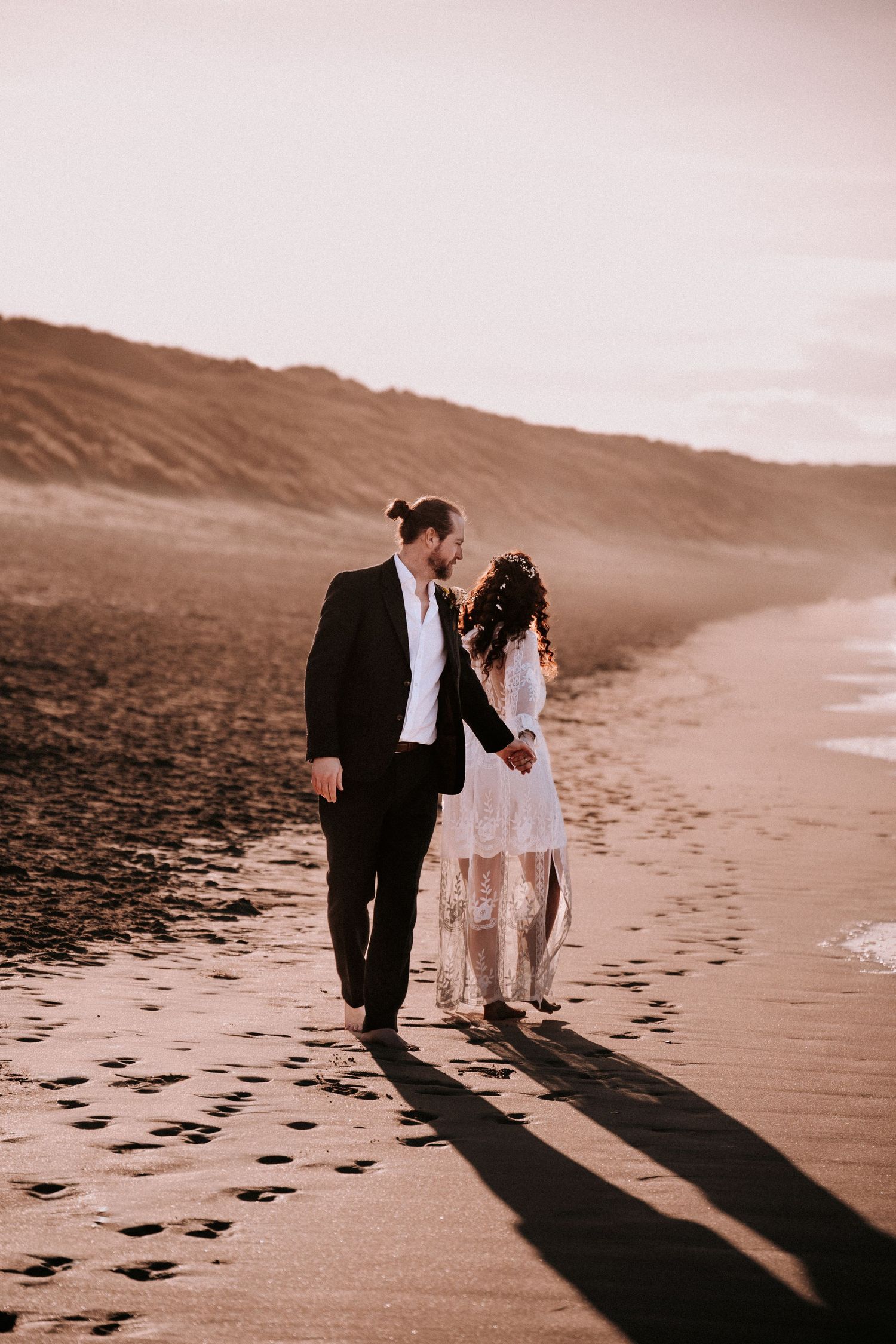 Bride and groom walk hand in hand along a sandy beach at golden hour, casting long shadows.