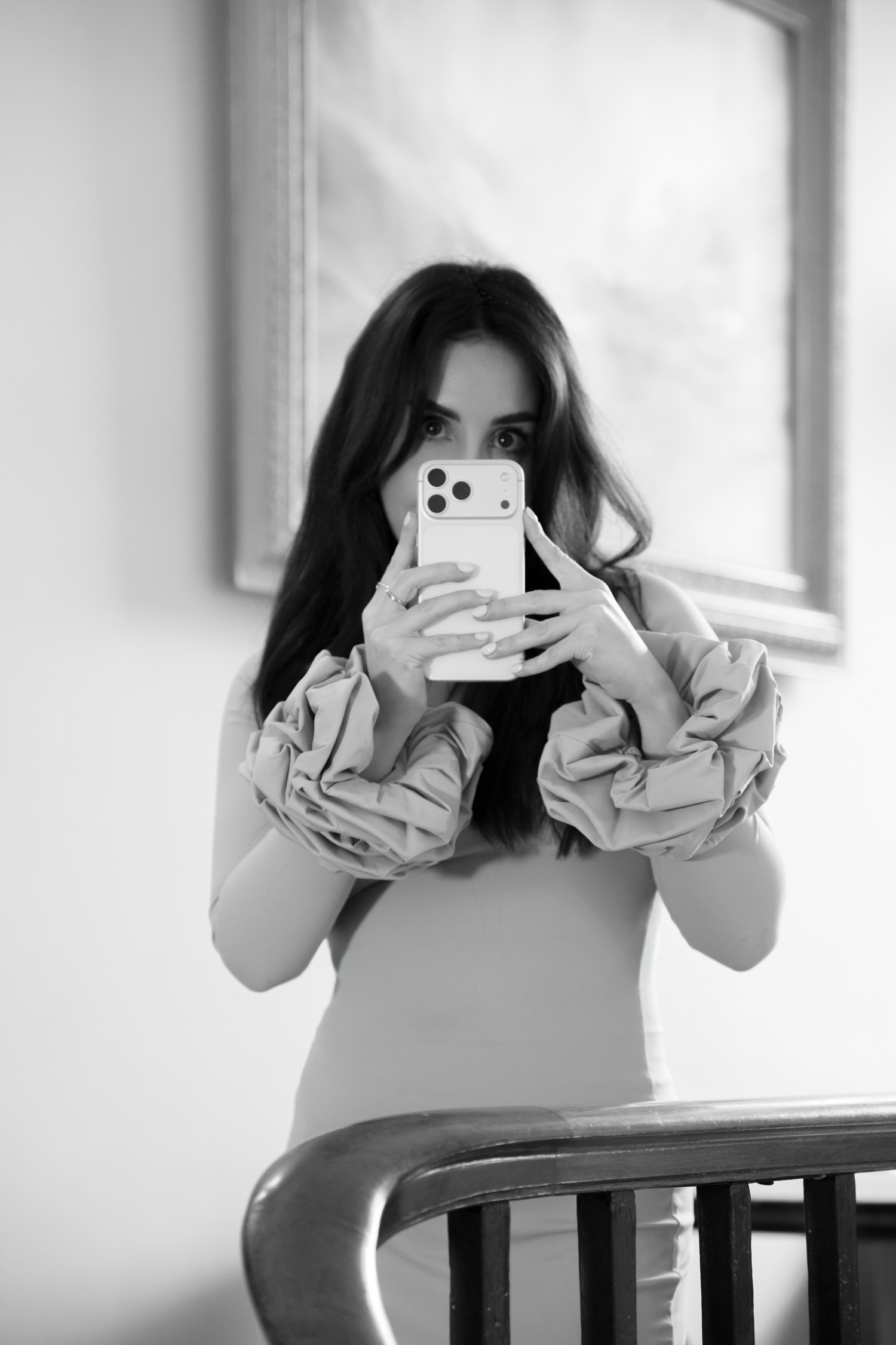Black and white mirror selfie of a woman with long dark hair wearing a dress with puffy sleeves, holding a smartphone.Content Creator.