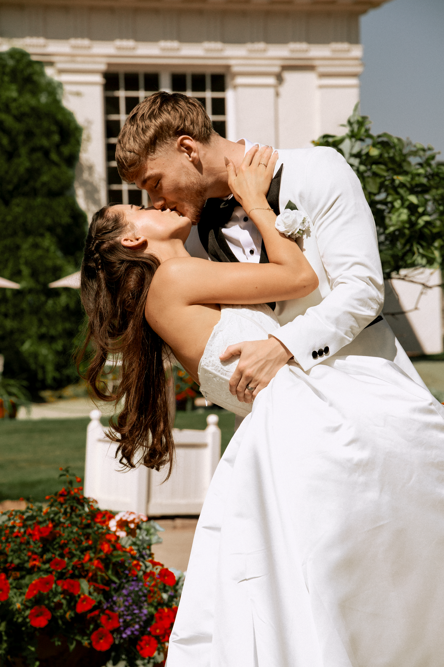 Bride and groom share a romantic dip kiss outside a white venue surrounded by red flowers.