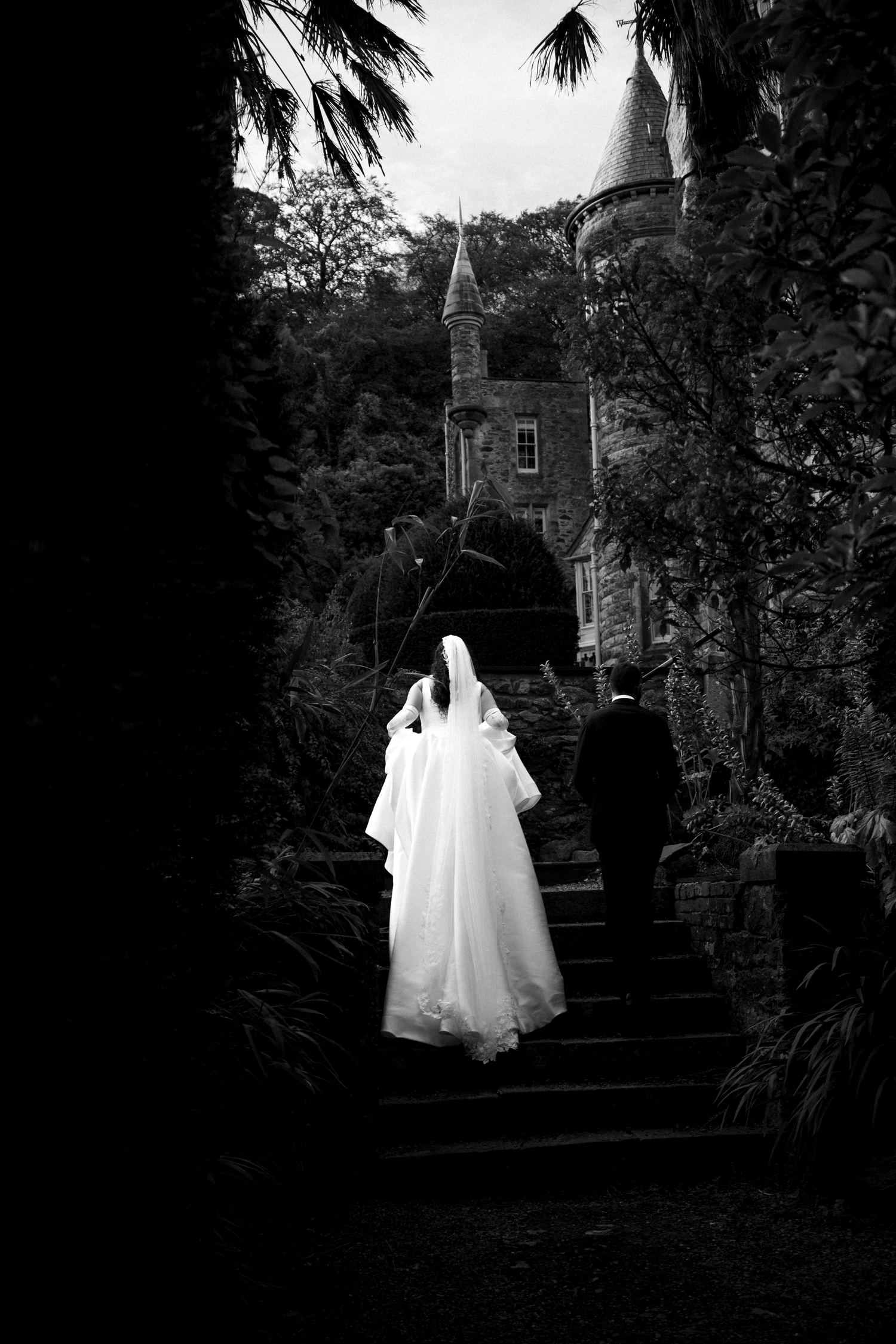 Black and white photo of a bride and groom ascending stone steps toward a castle surrounded by lush trees.