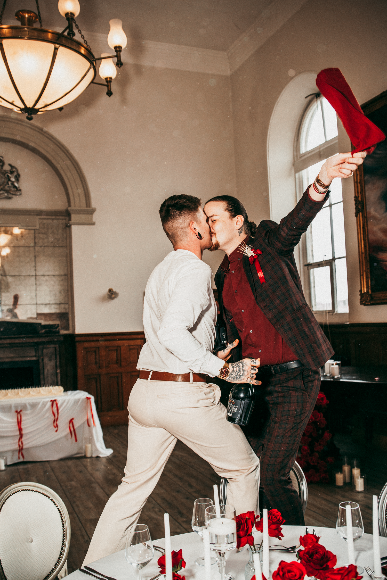 Two grooms kissing while standing on chair at wedding reception with red roses and candles.