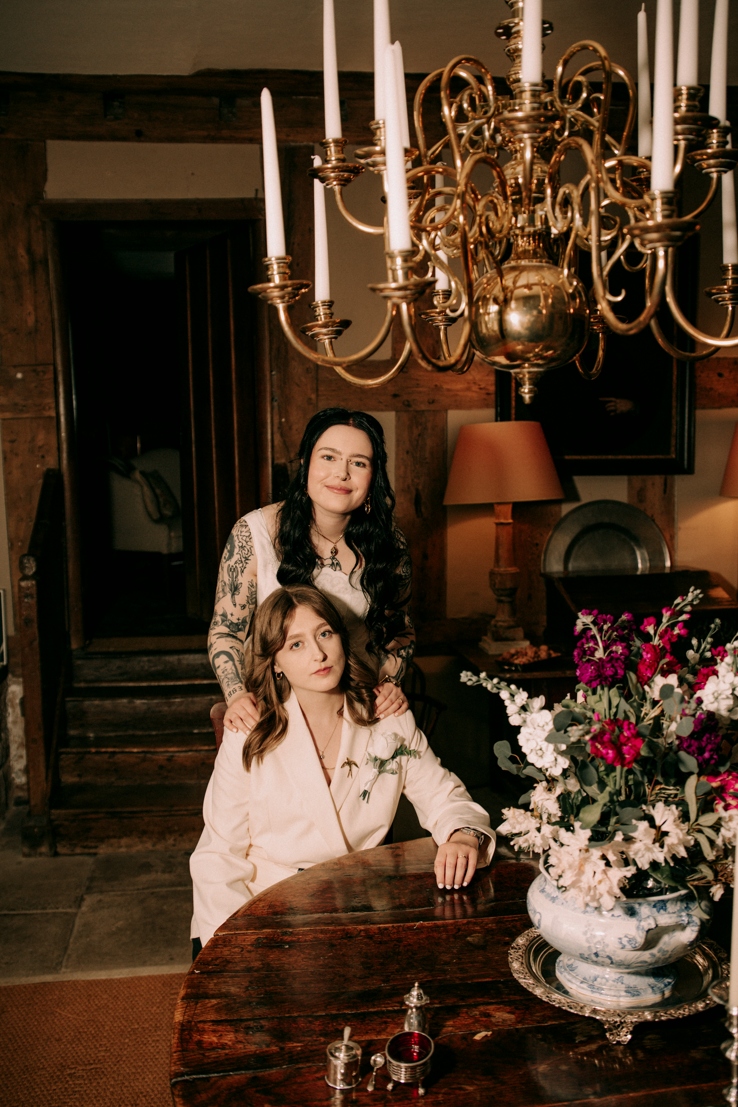 Two women in white robes posing at antique wooden table beneath gold chandelier with floral arrangement.