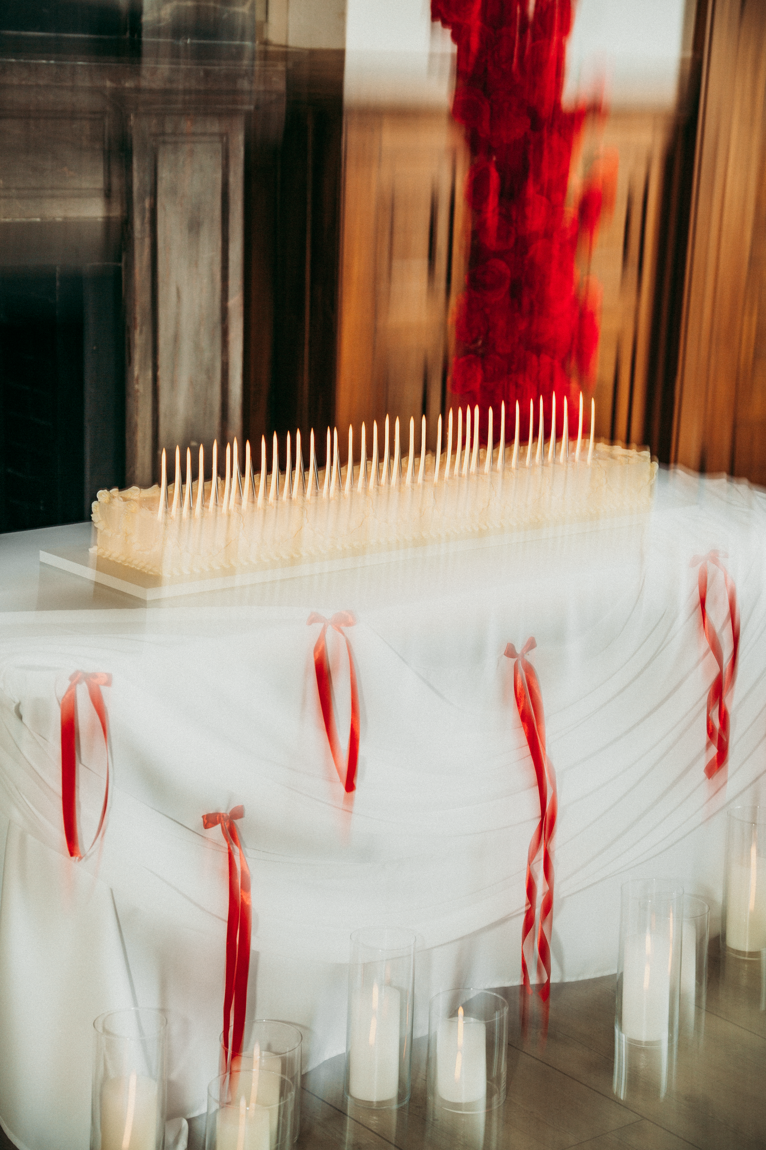 White wedding cake with tall candles and red ribbon bows on draped table with pillar candles.