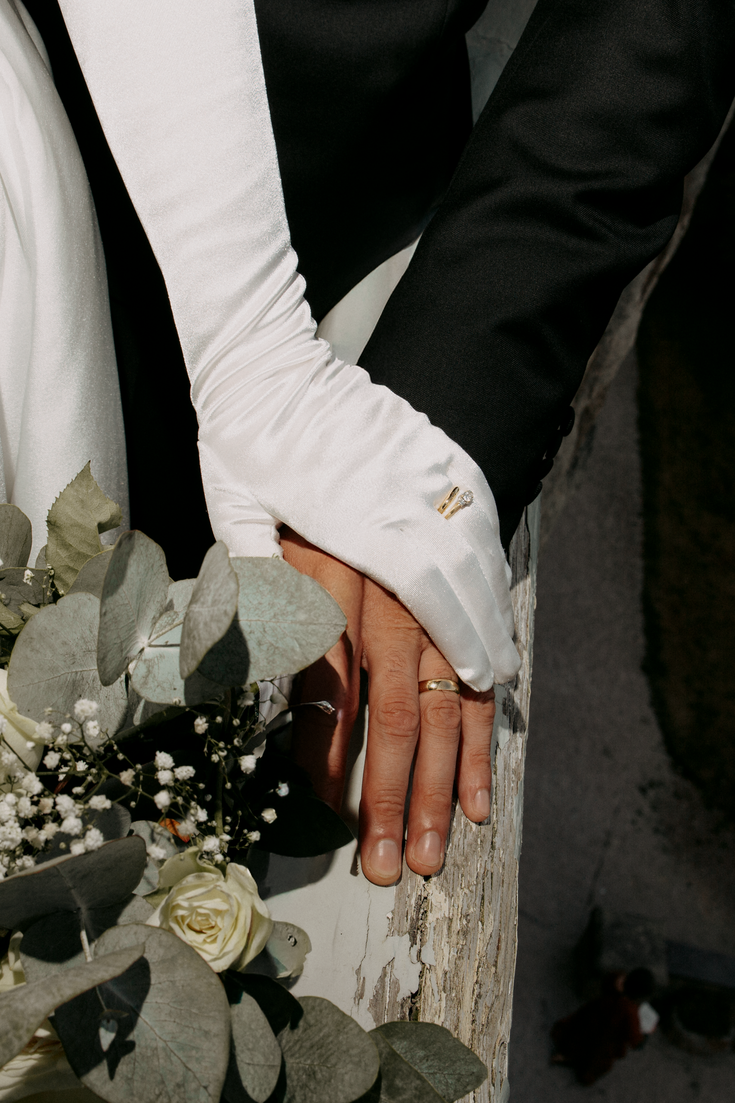 Bride and groom holding hands, showcasing wedding rings, white glove, and bouquet with roses and eucalyptus.