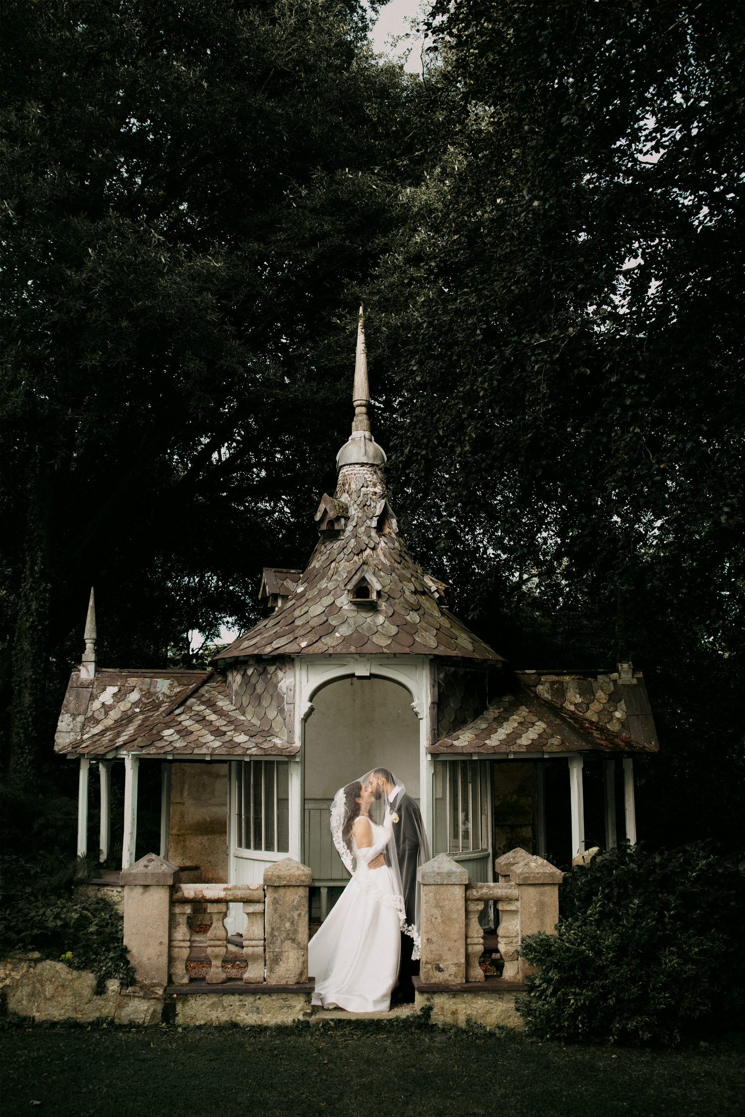 Bride and groom embracing in front of a rustic ornate gazebo surrounded by lush dark trees.