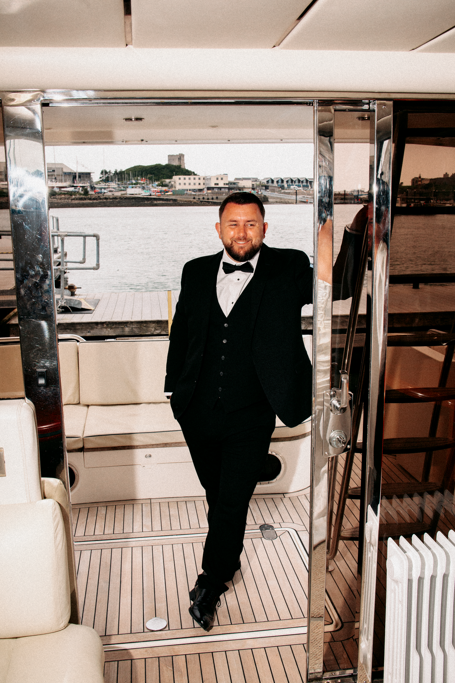 Dapper groom in black tuxedo leaning against yacht interior doorway with waterfront visible behind.