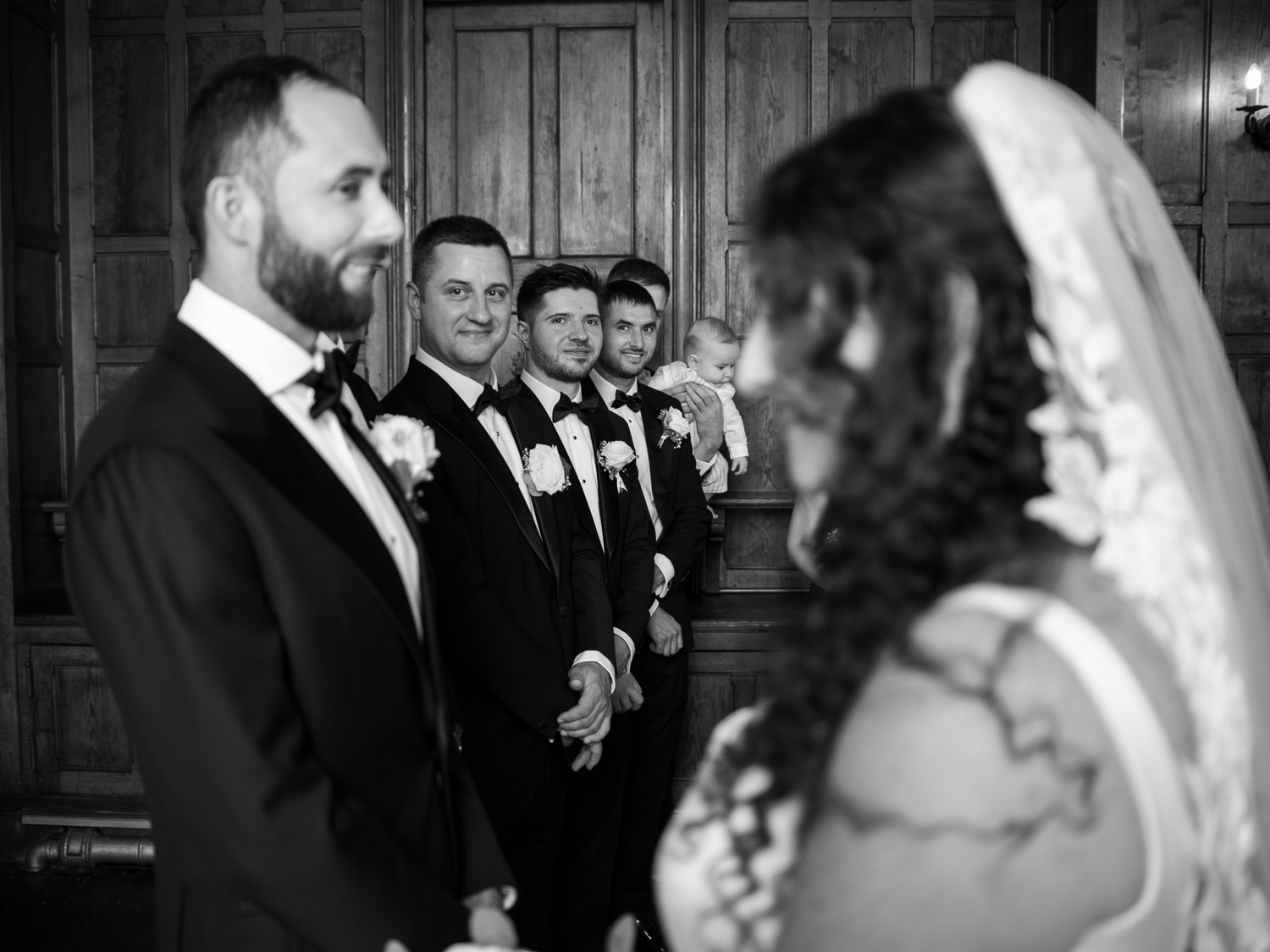 Black and white wedding photo of groom and groomsmen smiling at bride during ceremony.