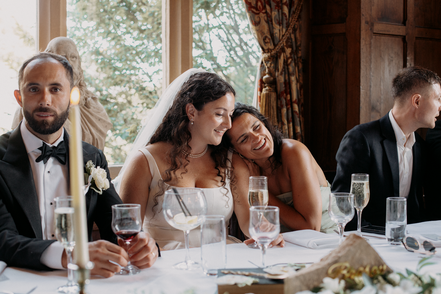 Two brides share a tender moment at their wedding reception table, smiling and leaning together with champagne glasses nearby.