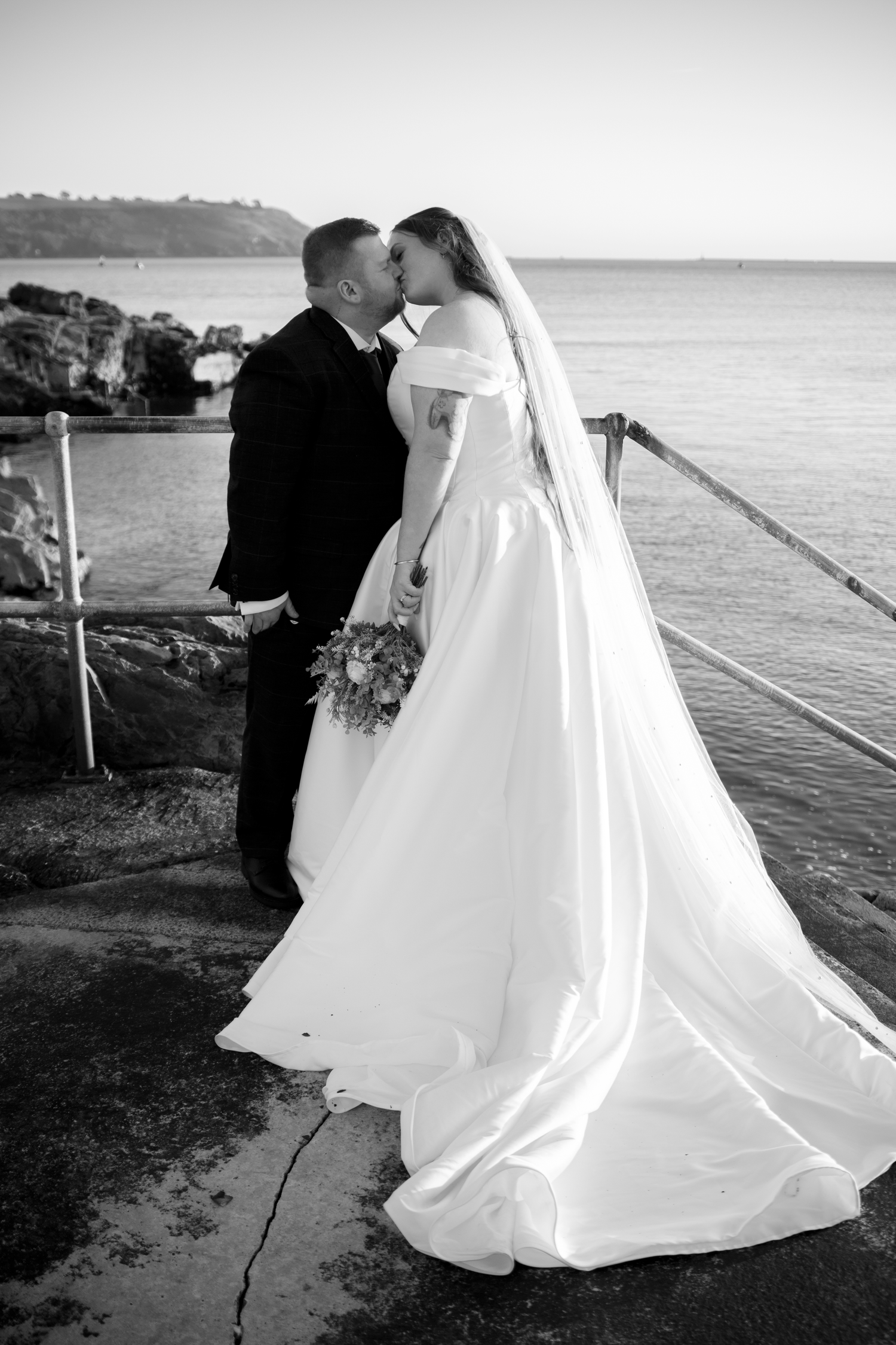 Black and white photo of bride and groom sharing a kiss by the ocean with flowing gown.