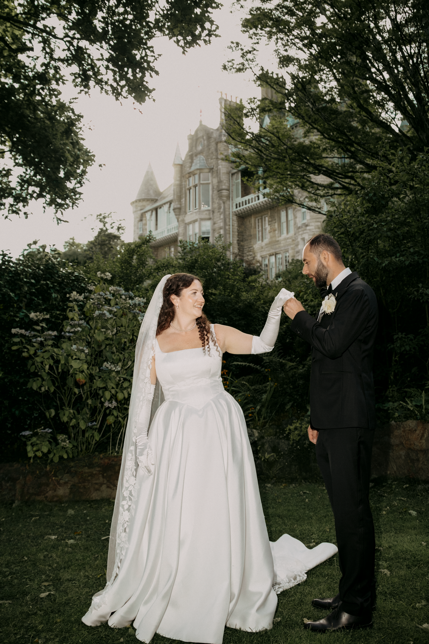 A bride and groom share a romantic moment in a lush garden with a grand castle backdrop on their wedding day.