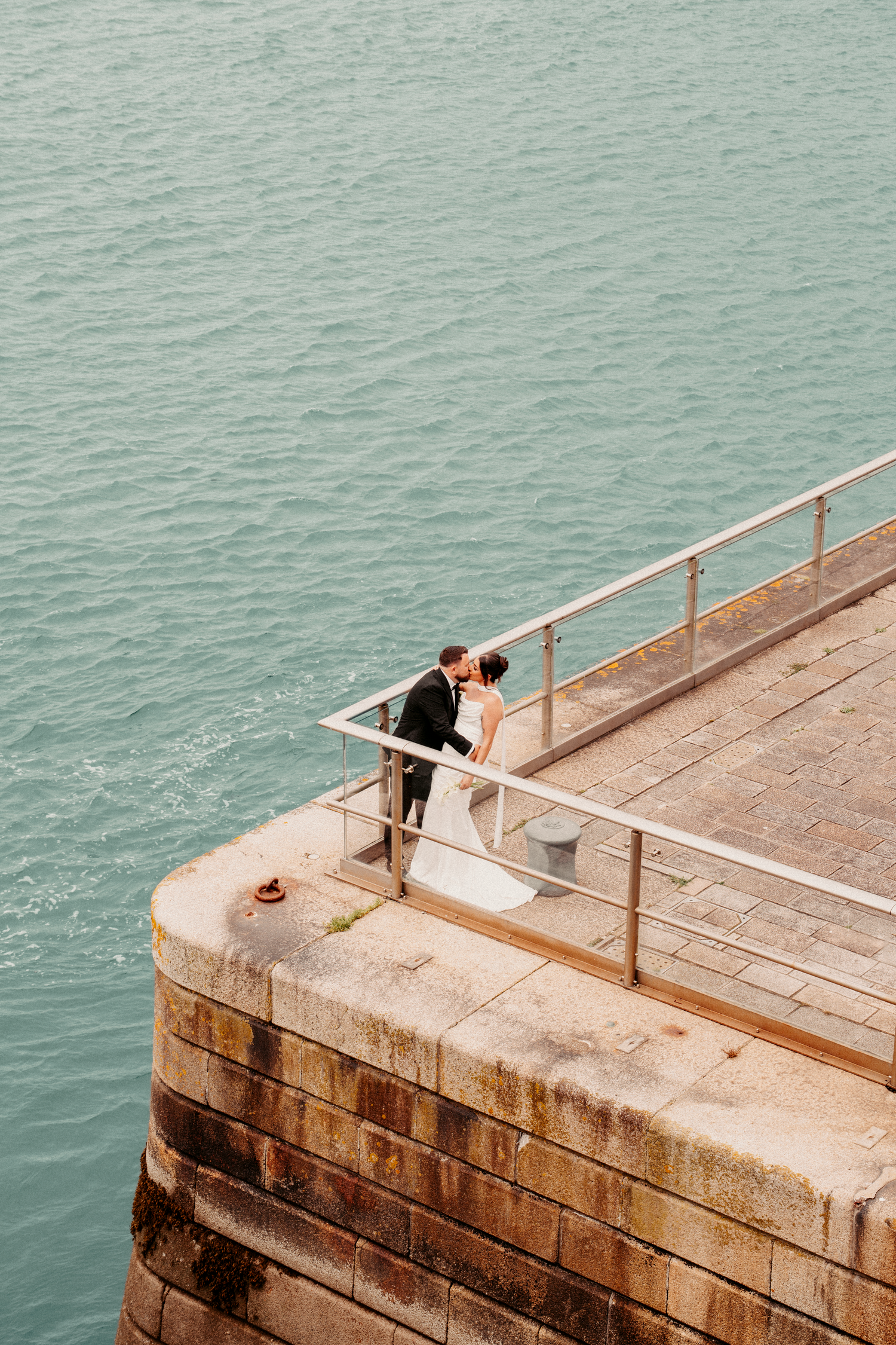 Bride and groom share a kiss on a stone pier overlooking turquoise water at their wedding.