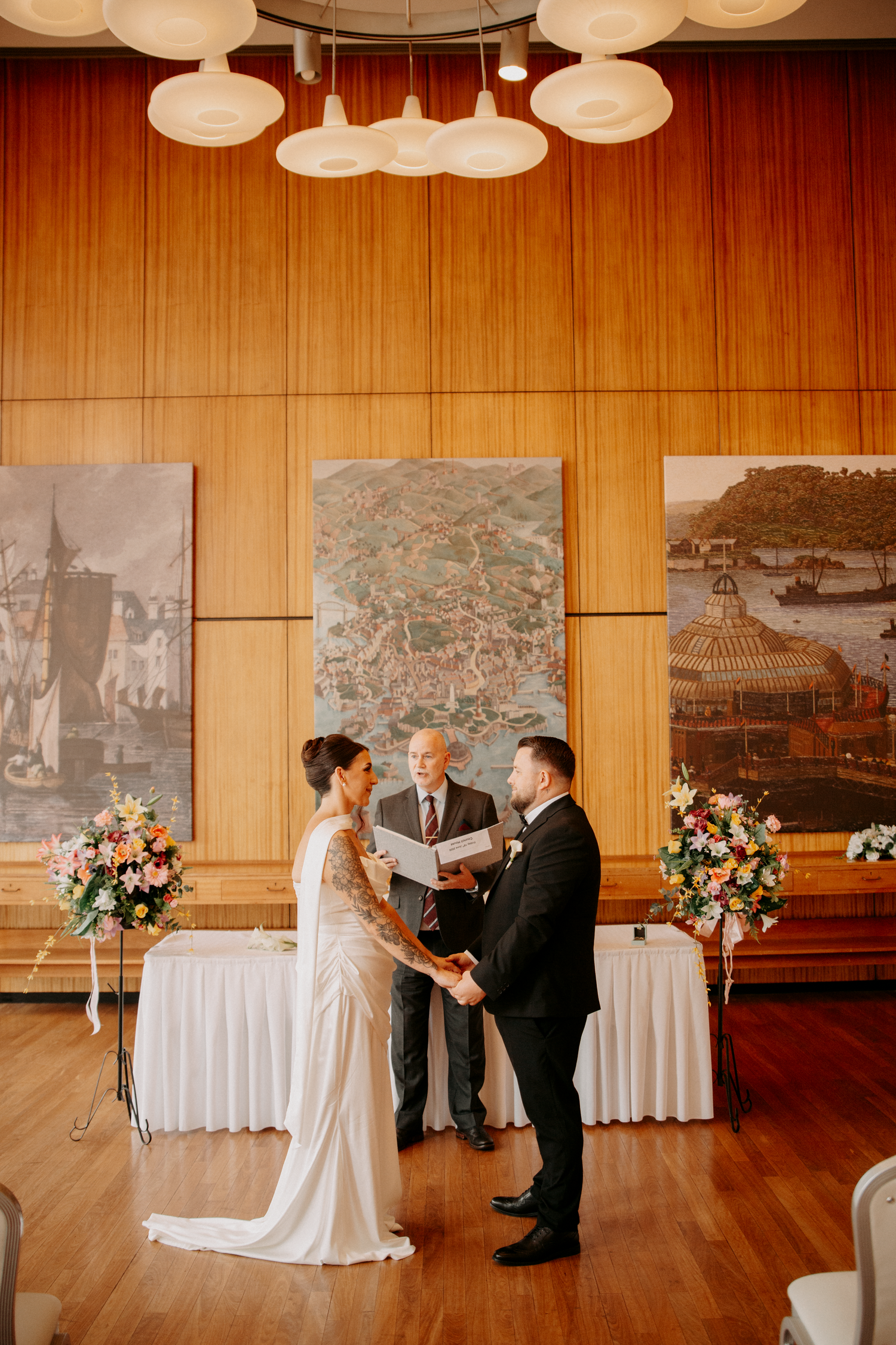 Bride and groom exchange vows before an officiant in a warmly lit wood-paneled wedding ceremony hall.