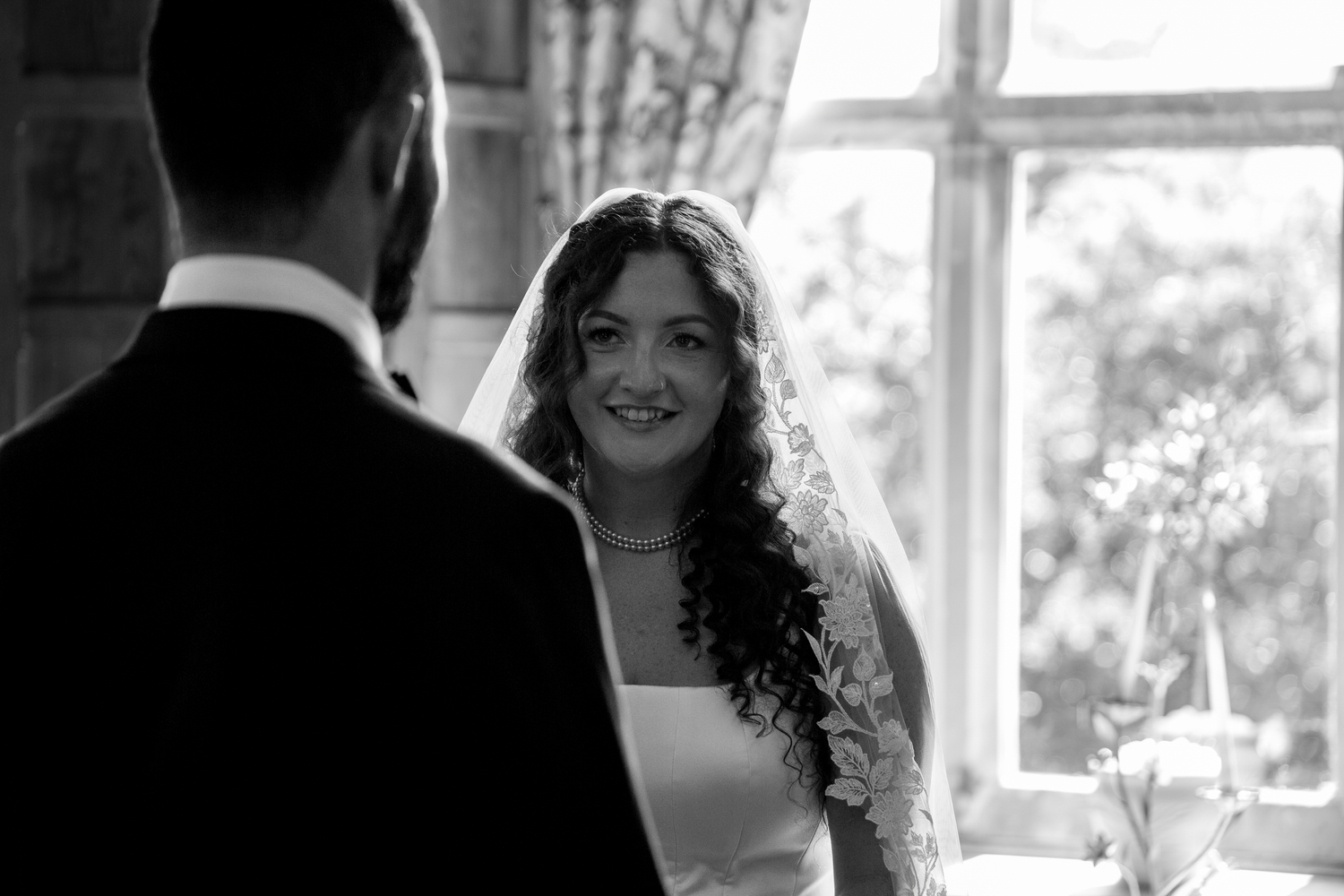 Black and white wedding photo of a smiling bride in veil facing groom indoors near large window.