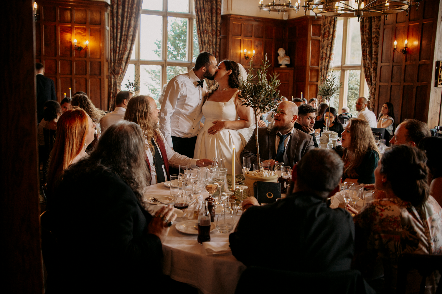 Bride and groom share a kiss at elegant wedding reception in wood-paneled venue with guests seated at candlelit tables.