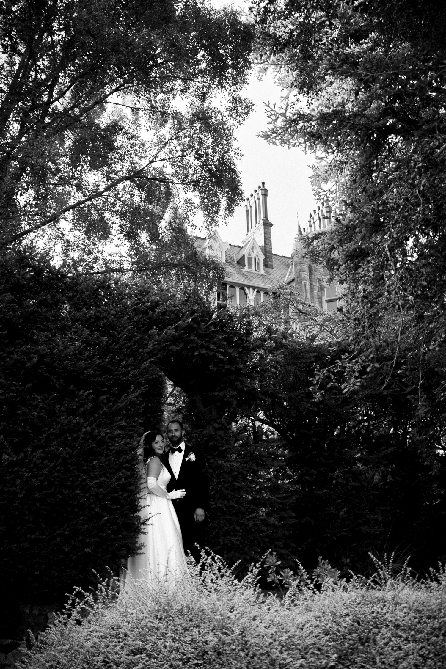 Black and white wedding photo of bride and groom embracing among lush trees with a historic manor in the background.