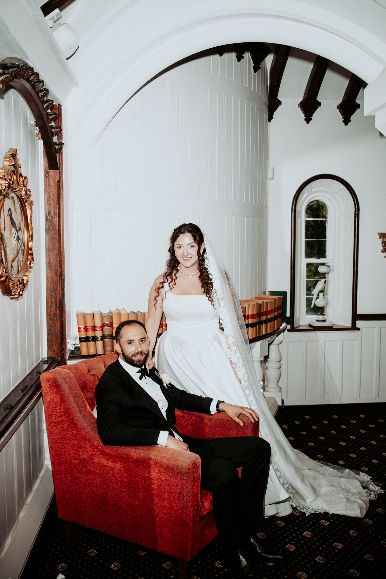 Bride in white gown stands behind groom in tuxedo seated in red velvet chair inside elegant venue.