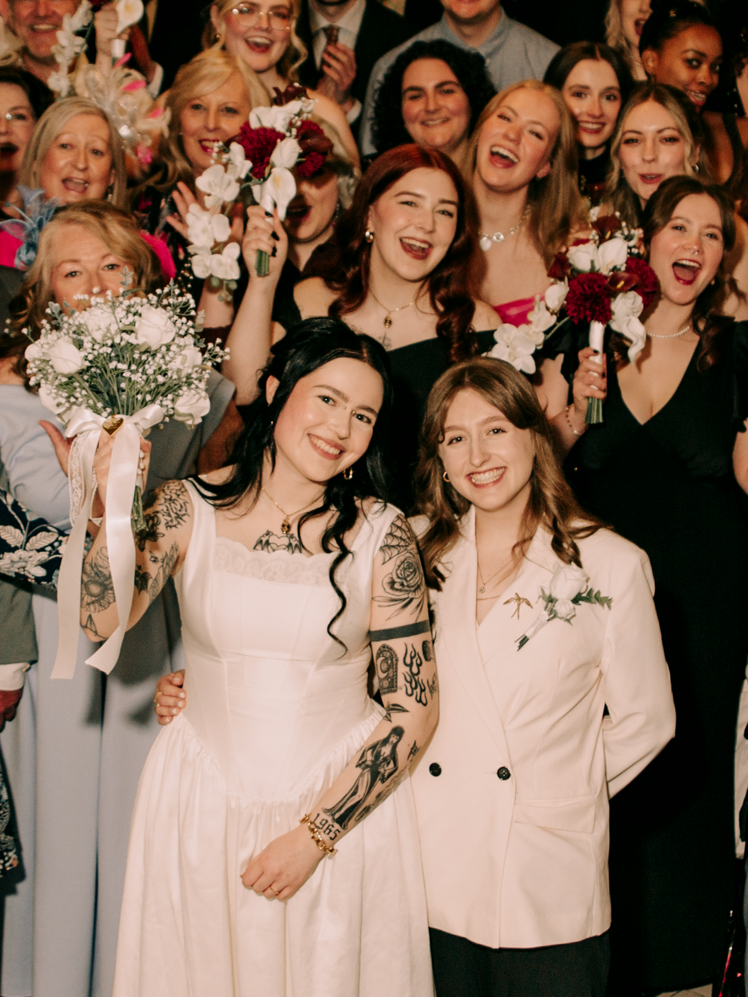 Two brides smiling with joyful wedding guests celebrating behind them, holding white and red bouquets.