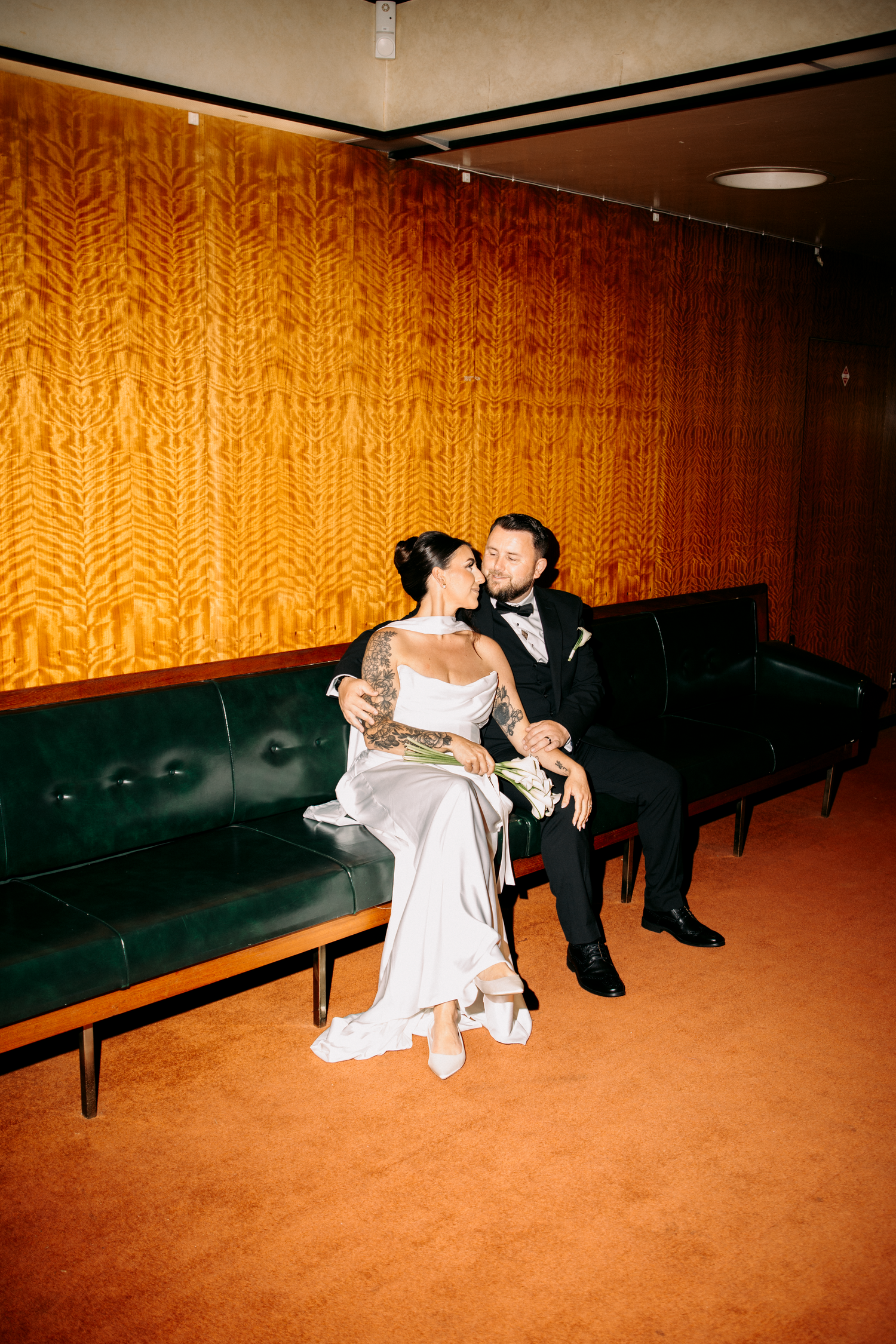 Bride and groom sit intimately on a green leather bench against a golden textured wall.
