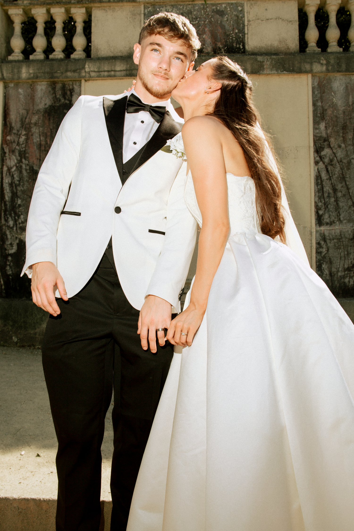 Bride in white strapless gown kissing groom in white tuxedo jacket on cheek while holding hands outdoors.