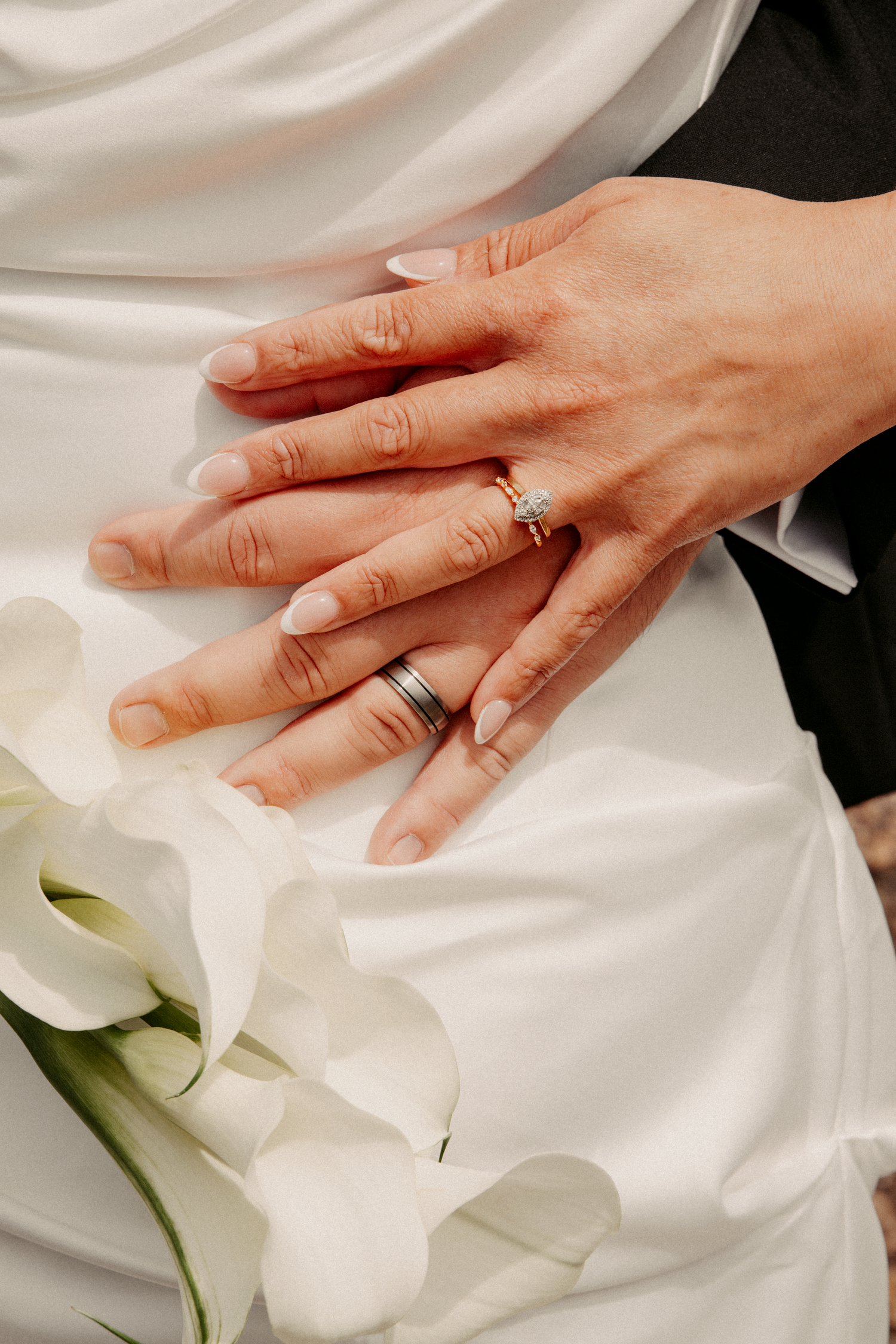 Close-up of newlyweds hands showing wedding rings resting on white bridal gown with calla lilies.