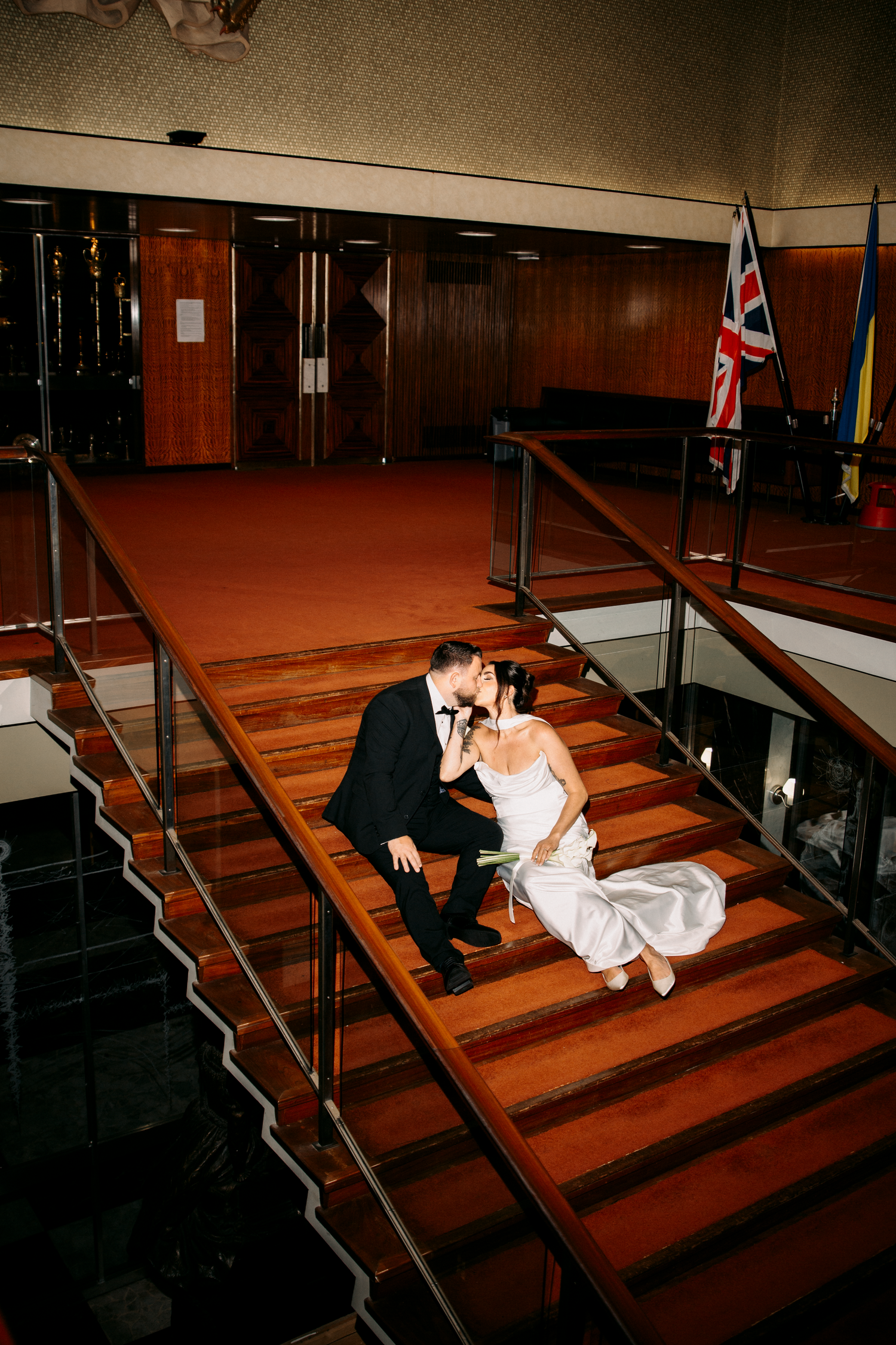 Bride and groom share a kiss seated on grand red carpeted staircase inside elegant venue.