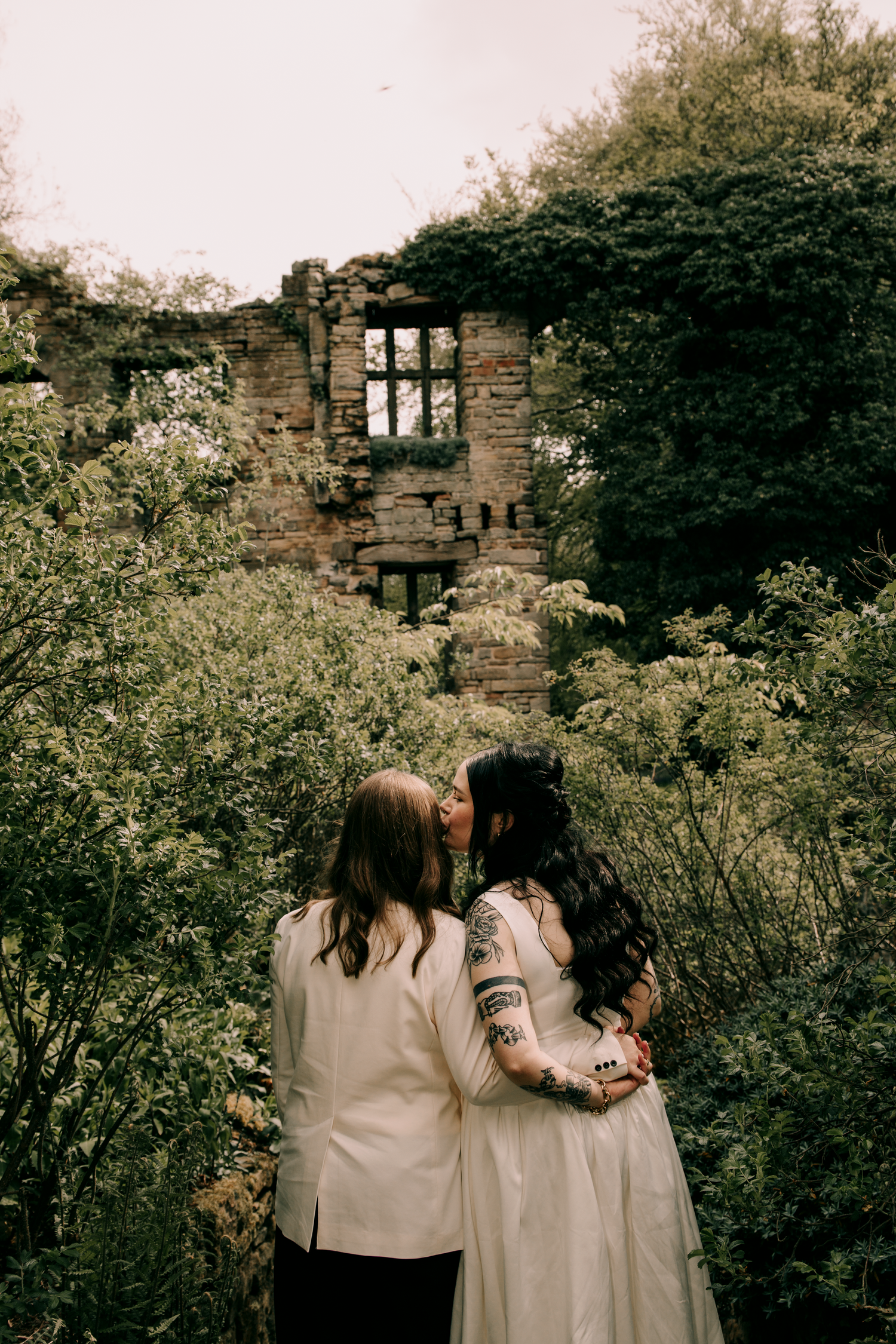 Two brides sharing a romantic kiss surrounded by overgrown ruins and lush greenery on their wedding day.