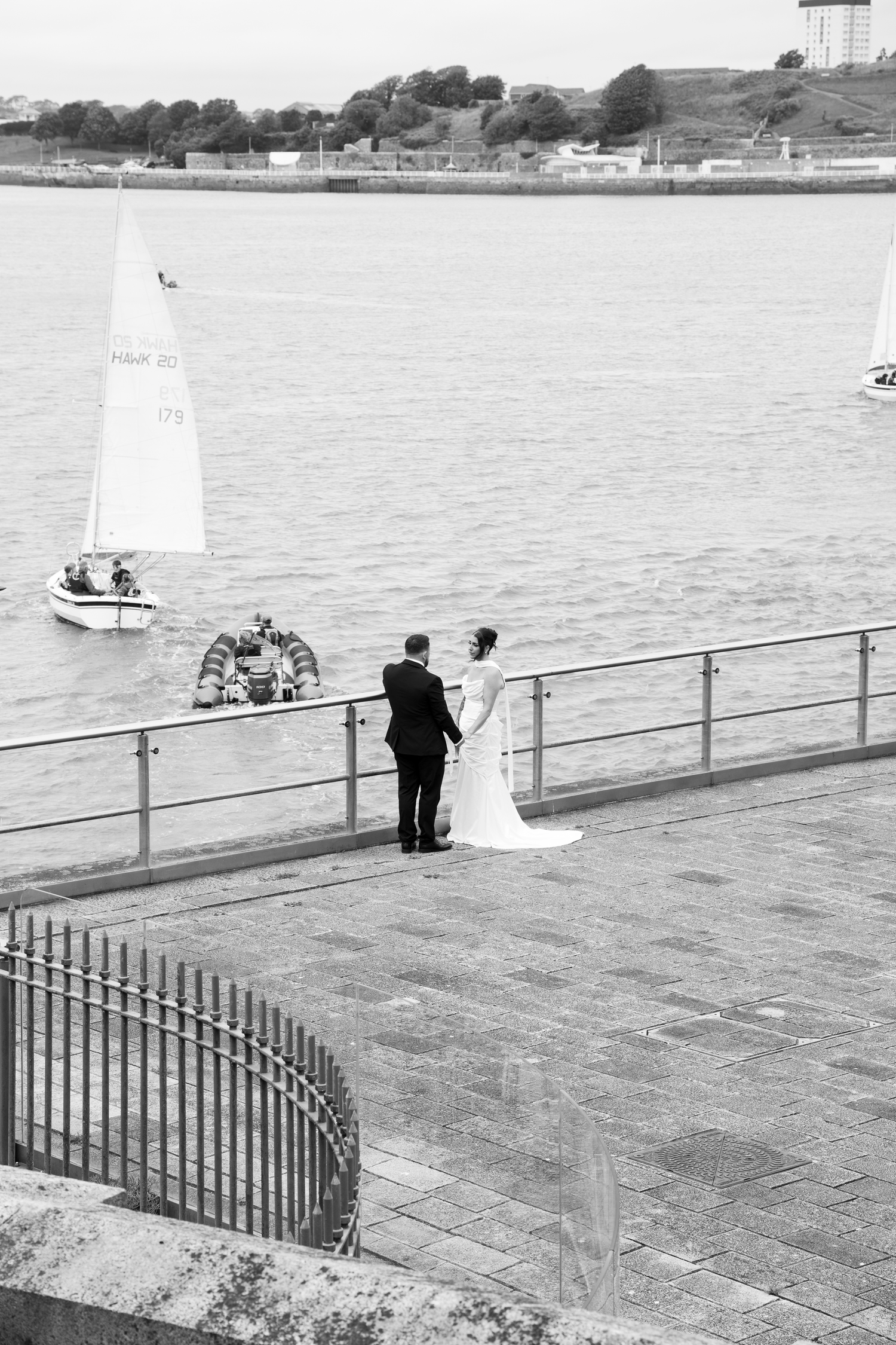 Bride and groom stand by waterfront railing overlooking sailboats on a calm harbor in black and white.