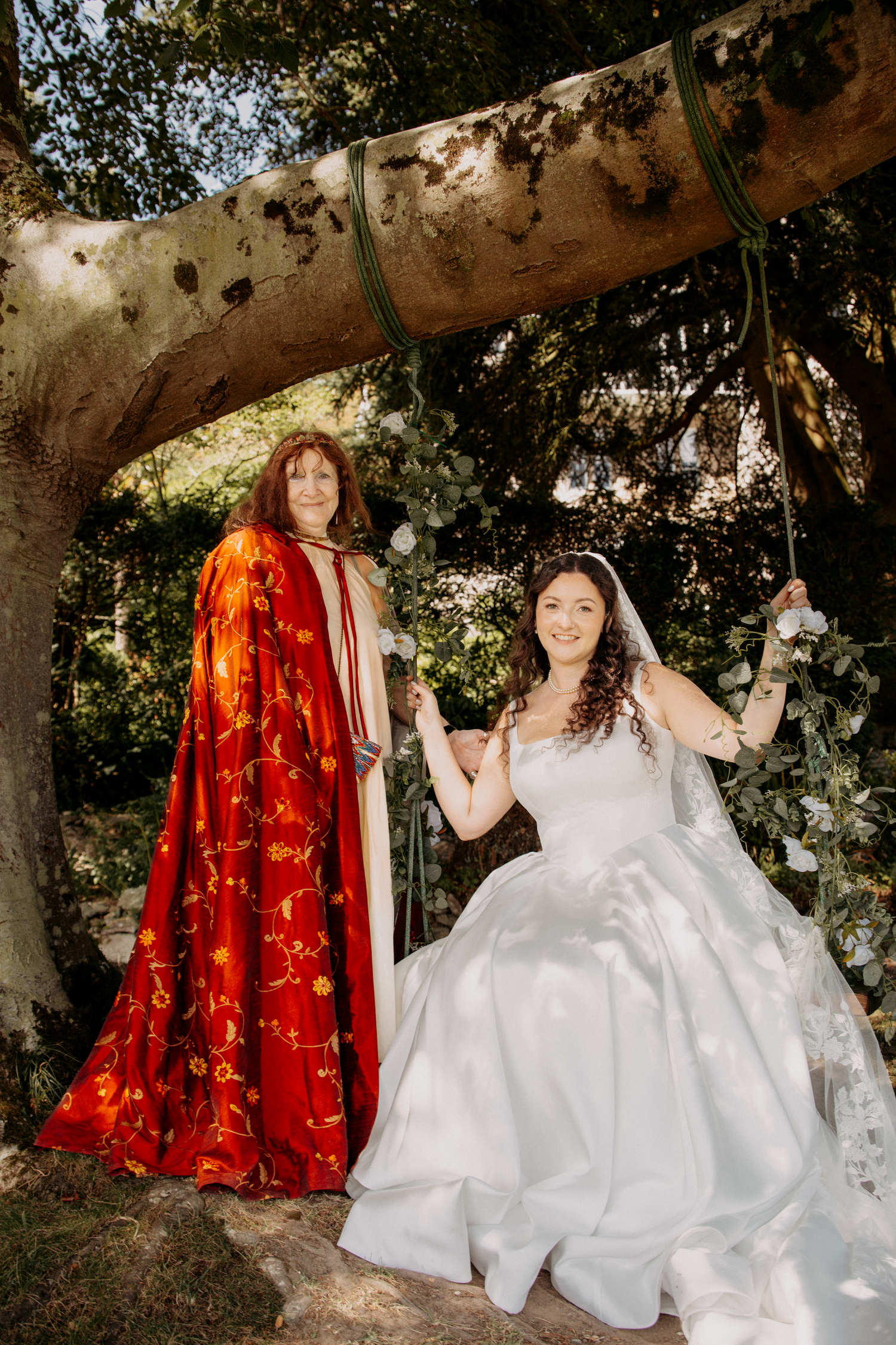 Bride in white gown sits on tree swing beside woman in red embroidered cloak at outdoor wedding ceremony.