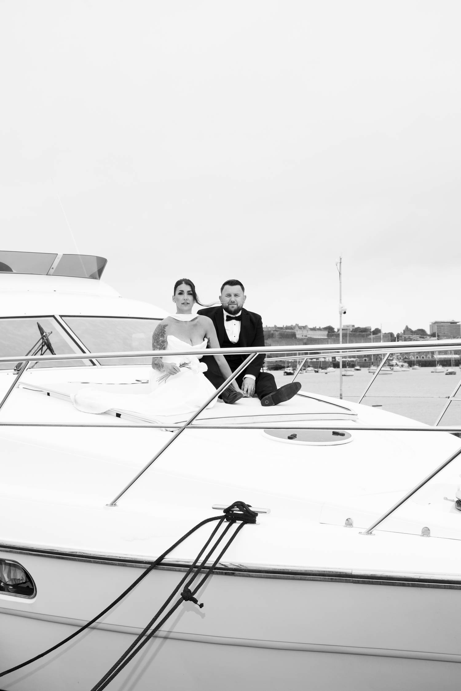 Black and white image of elegant bride and groom seated together on the bow of a harbor yacht.