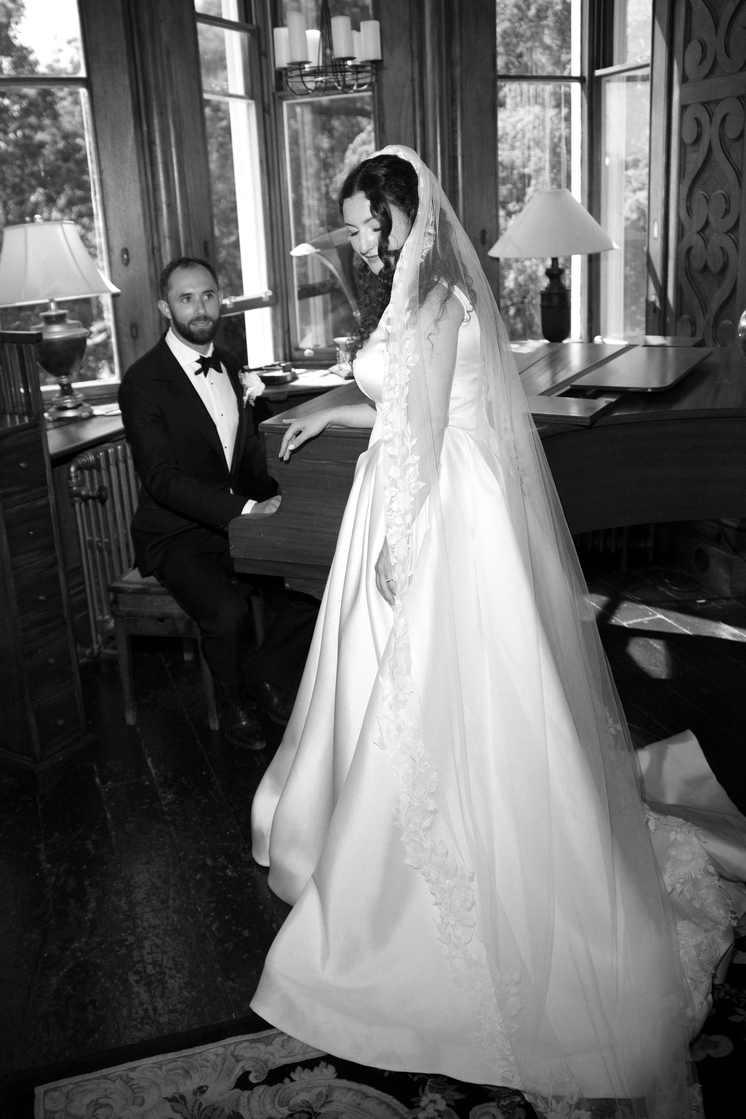 Black and white wedding photo of bride in elegant gown with veil and groom in tuxedo near a grand piano.