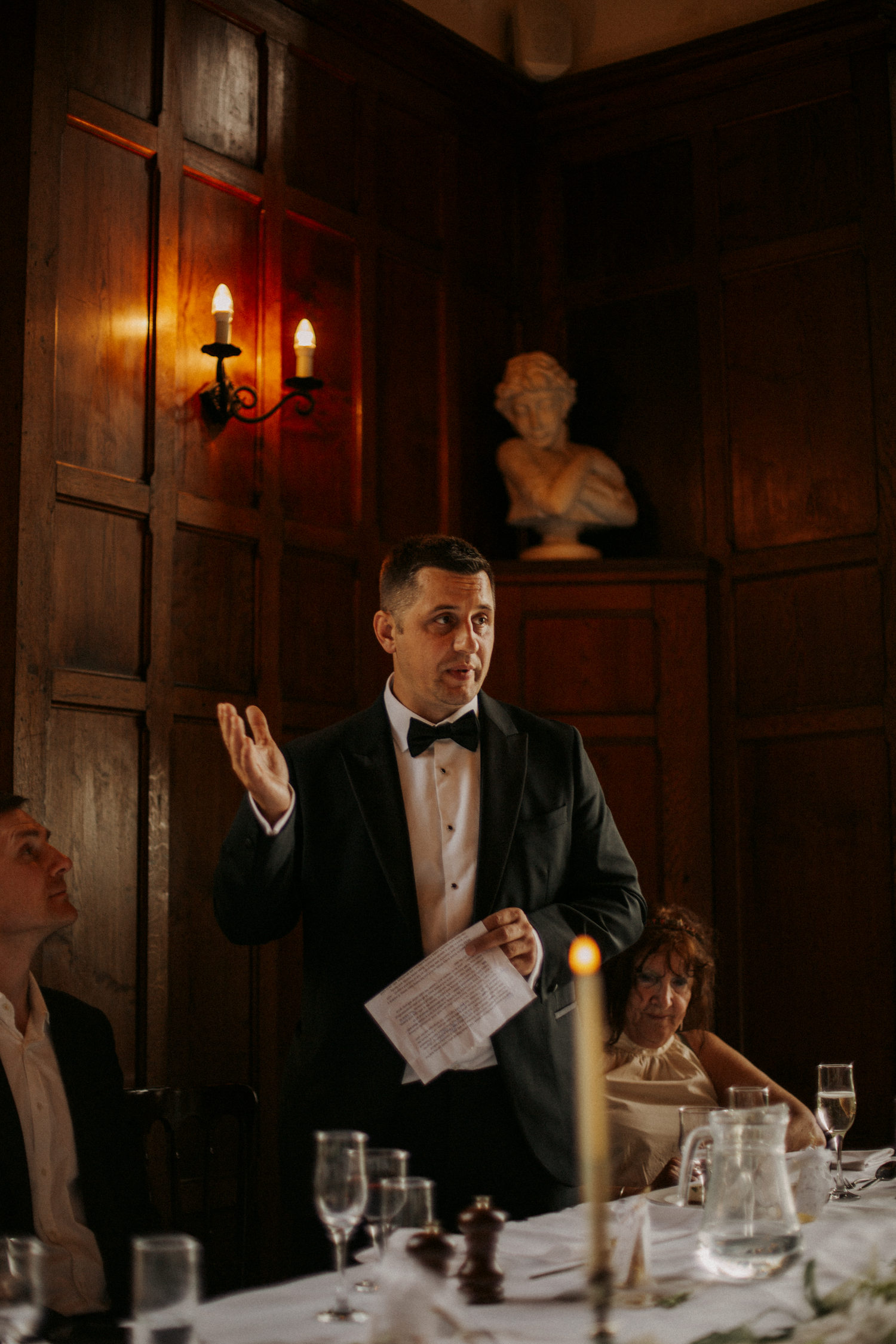 A man in a tuxedo delivers a speech at an elegant wedding dinner, holding notes beside candlelit tables.