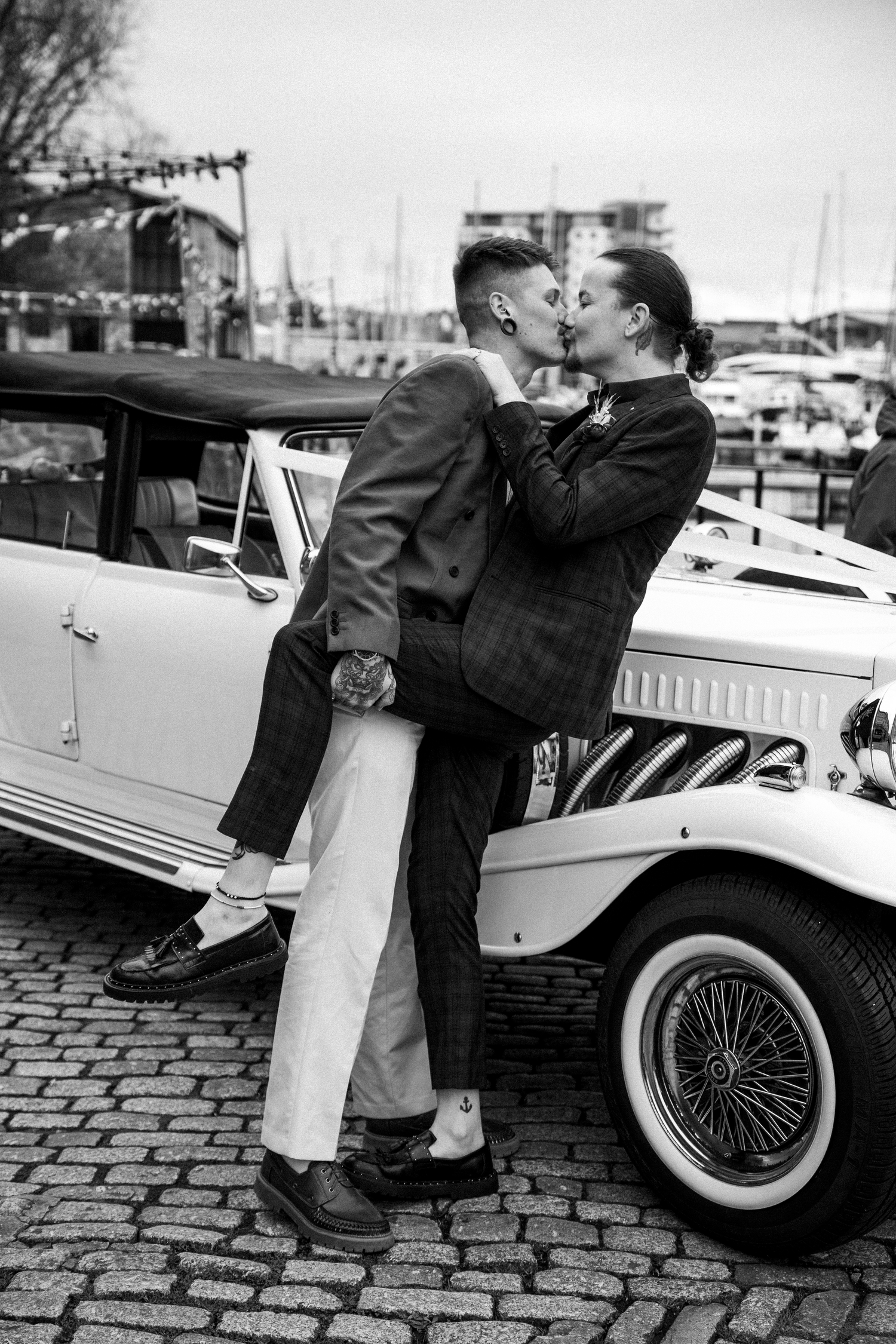 Two grooms share a romantic kiss leaning against a vintage white car at a harbor in black and white.