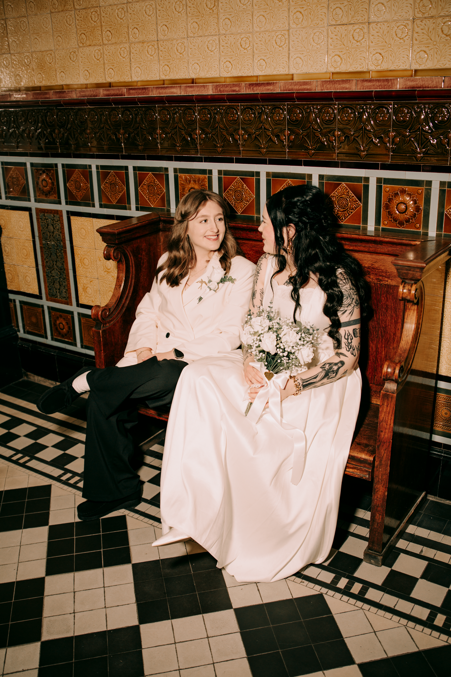 Two brides in white wedding attire sitting together on ornate wooden bench with decorative tiled backdrop.