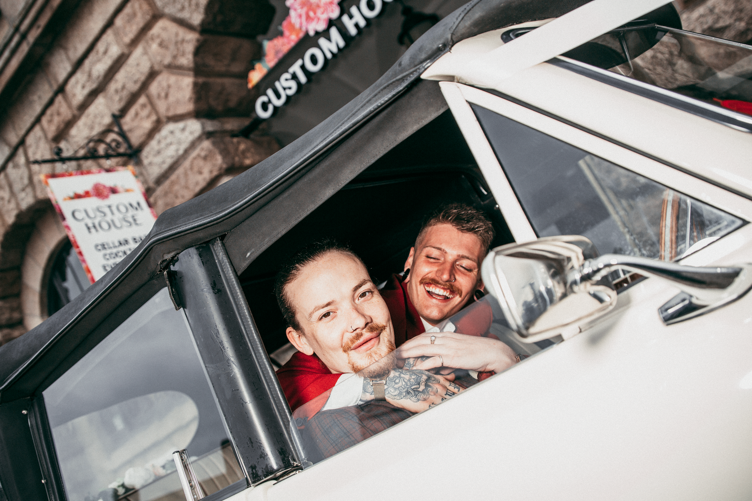 Two happy grooms smile out of a vintage white wedding car window outside Custom House venue.