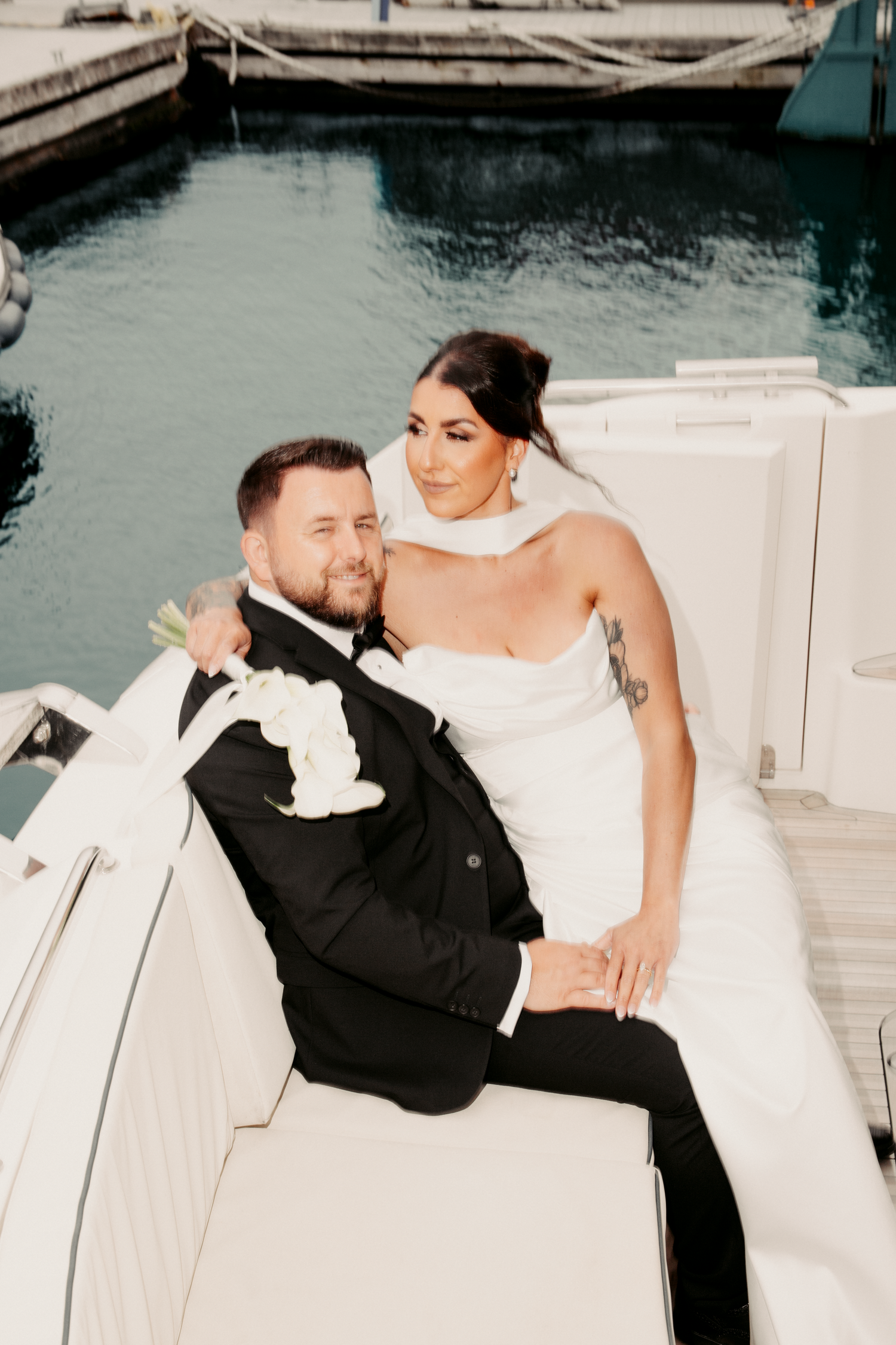 Bride and groom sitting together on a luxury yacht, bride in strapless white gown with tattoos, groom in black tuxedo.