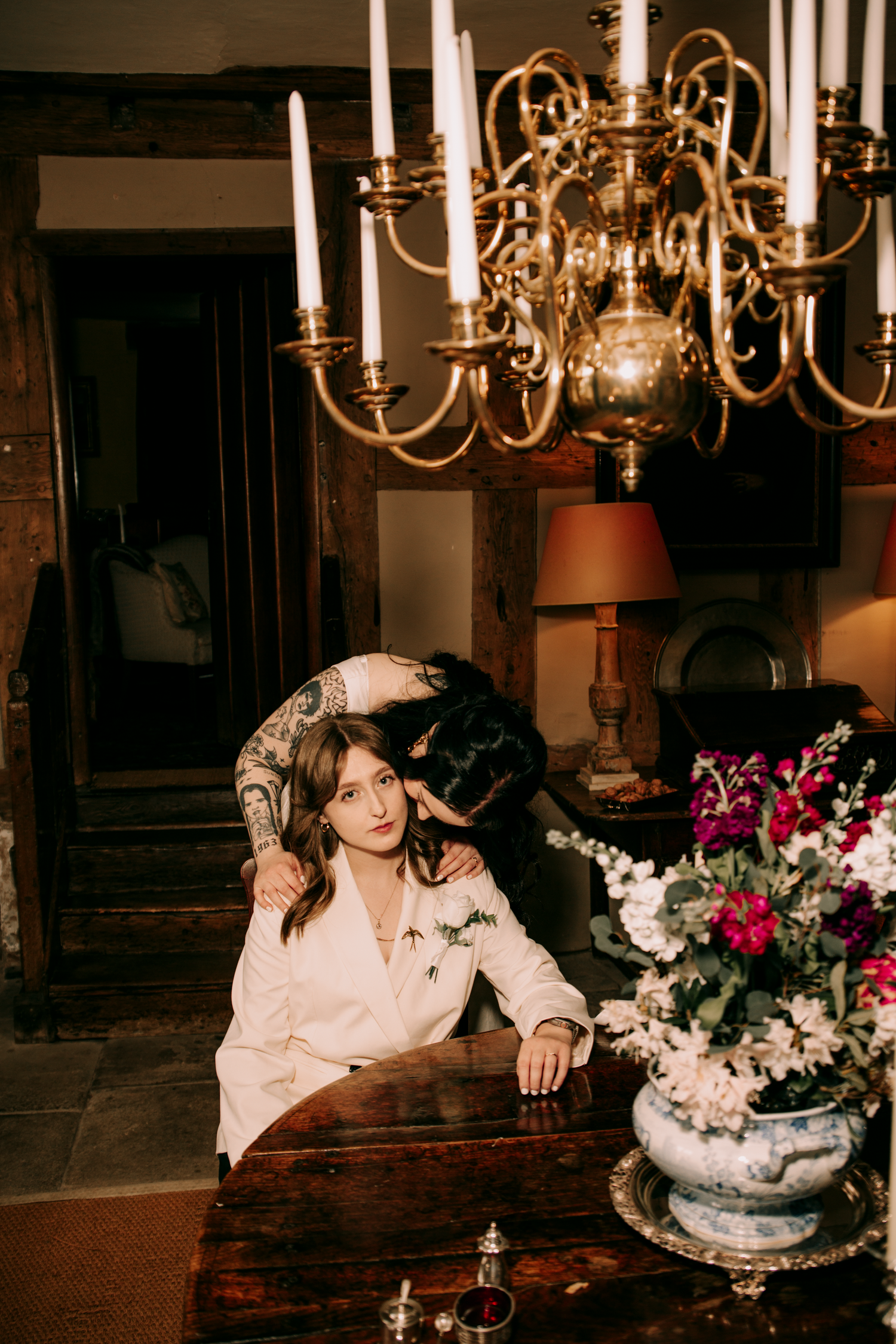 A bride sits at an antique table while her partner lovingly embraces her beneath a golden chandelier.
