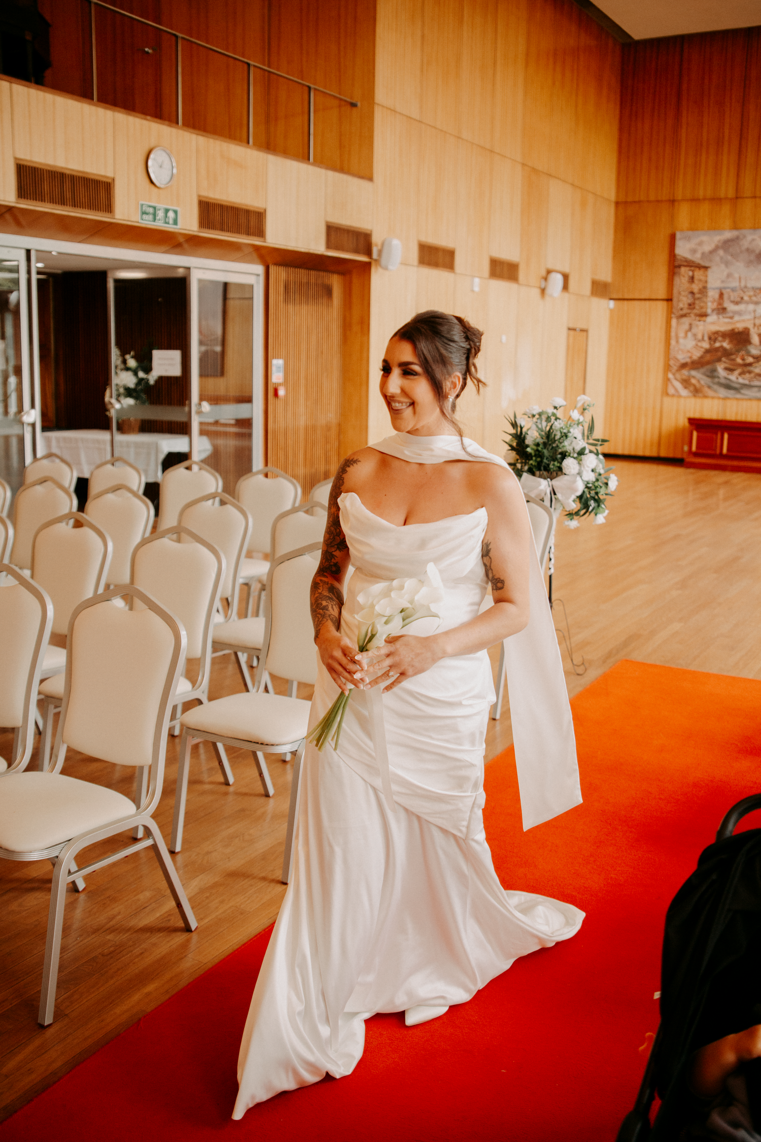 Elegant bride in a sleek white satin gown walks the red carpet aisle holding white calla lilies.