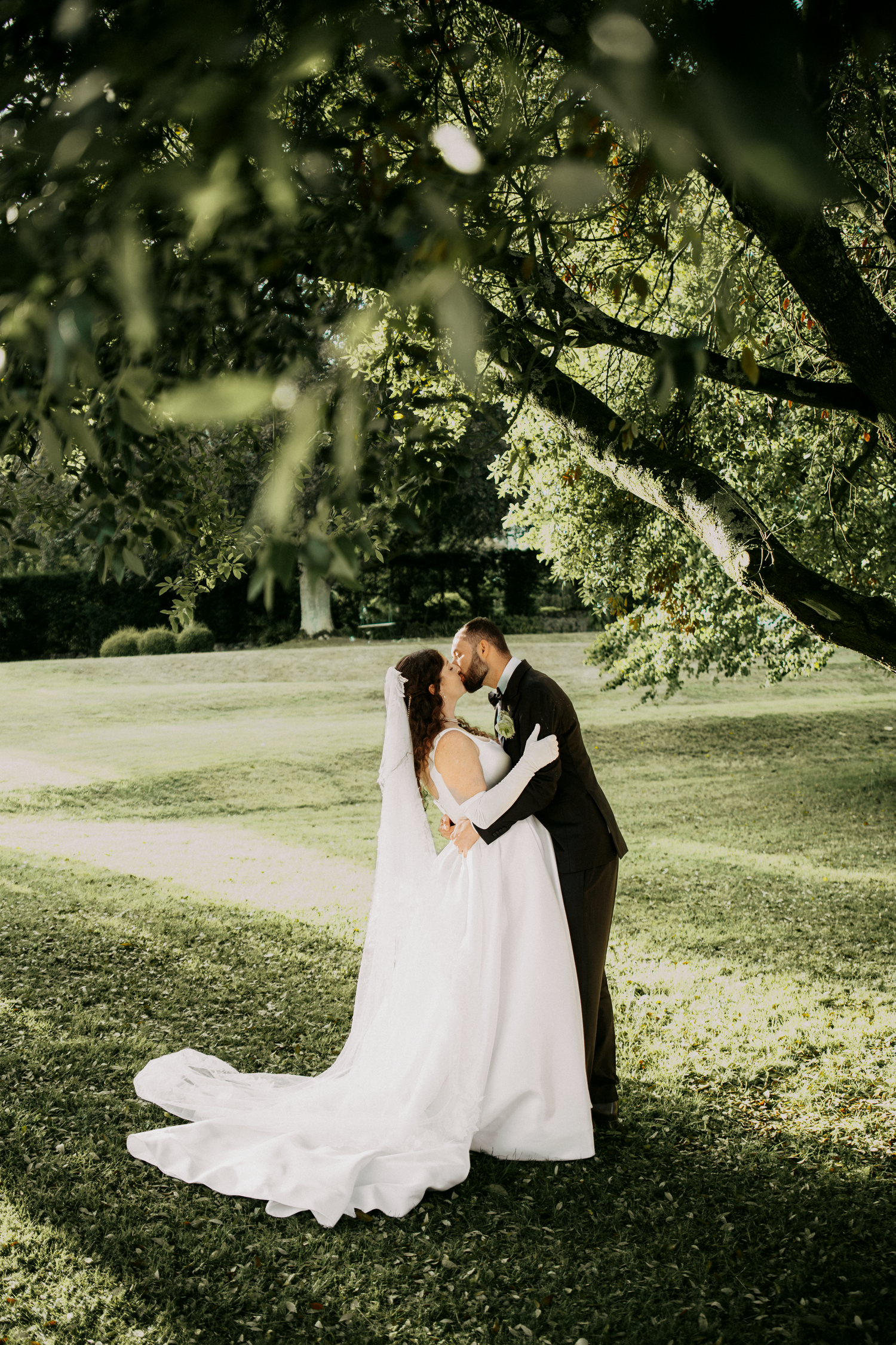A bride and groom share a romantic kiss under lush green trees on a sunlit lawn, her white gown trailing elegantly.
