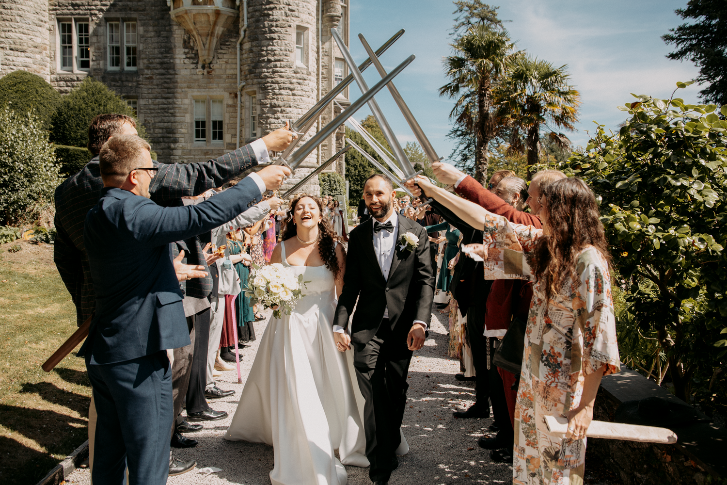 Bride and groom walk through a sword arch at an outdoor castle wedding ceremony on a sunny day.