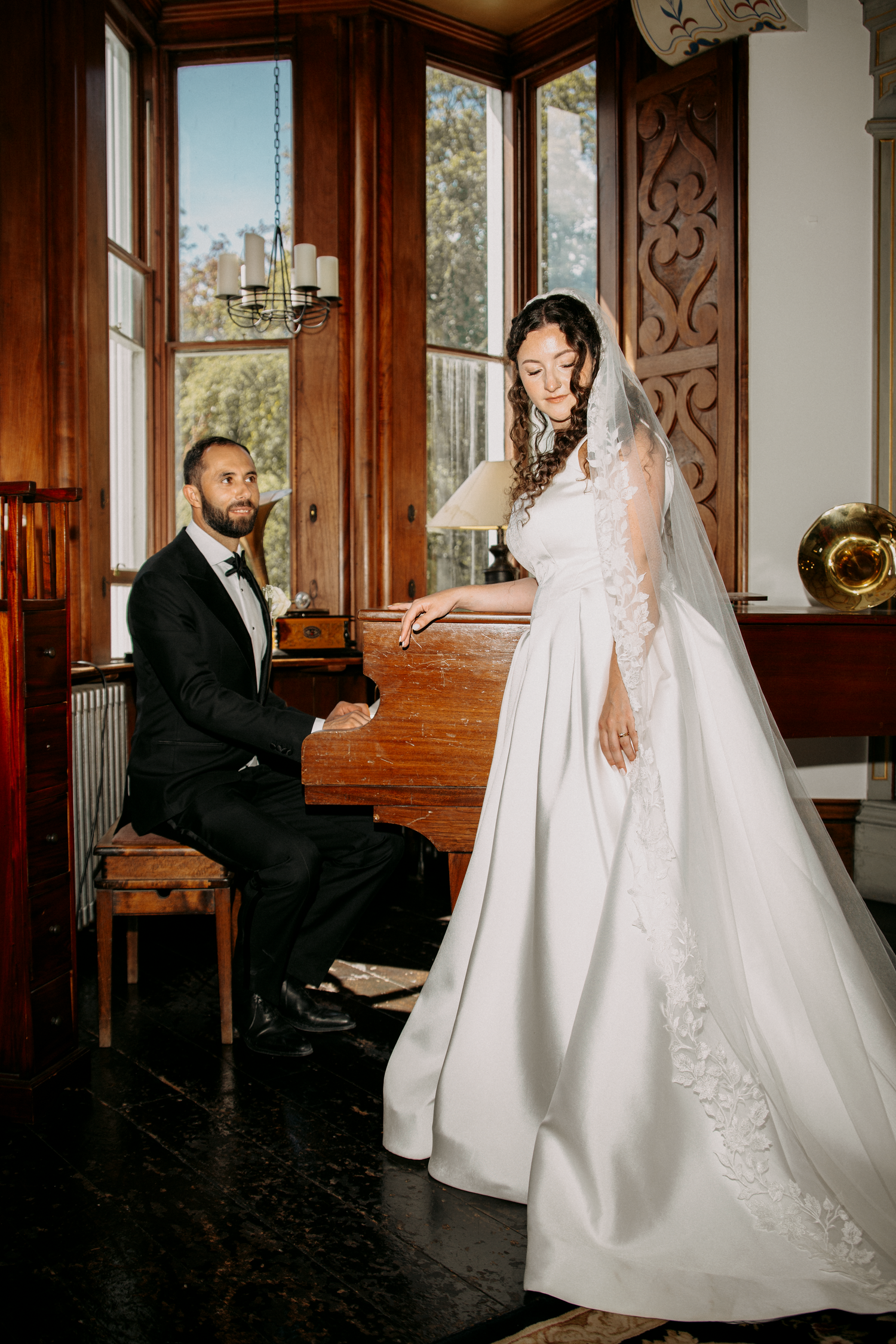 Groom in black tuxedo plays piano while bride in white lace wedding gown and veil stands nearby in elegant wood-paneled room.