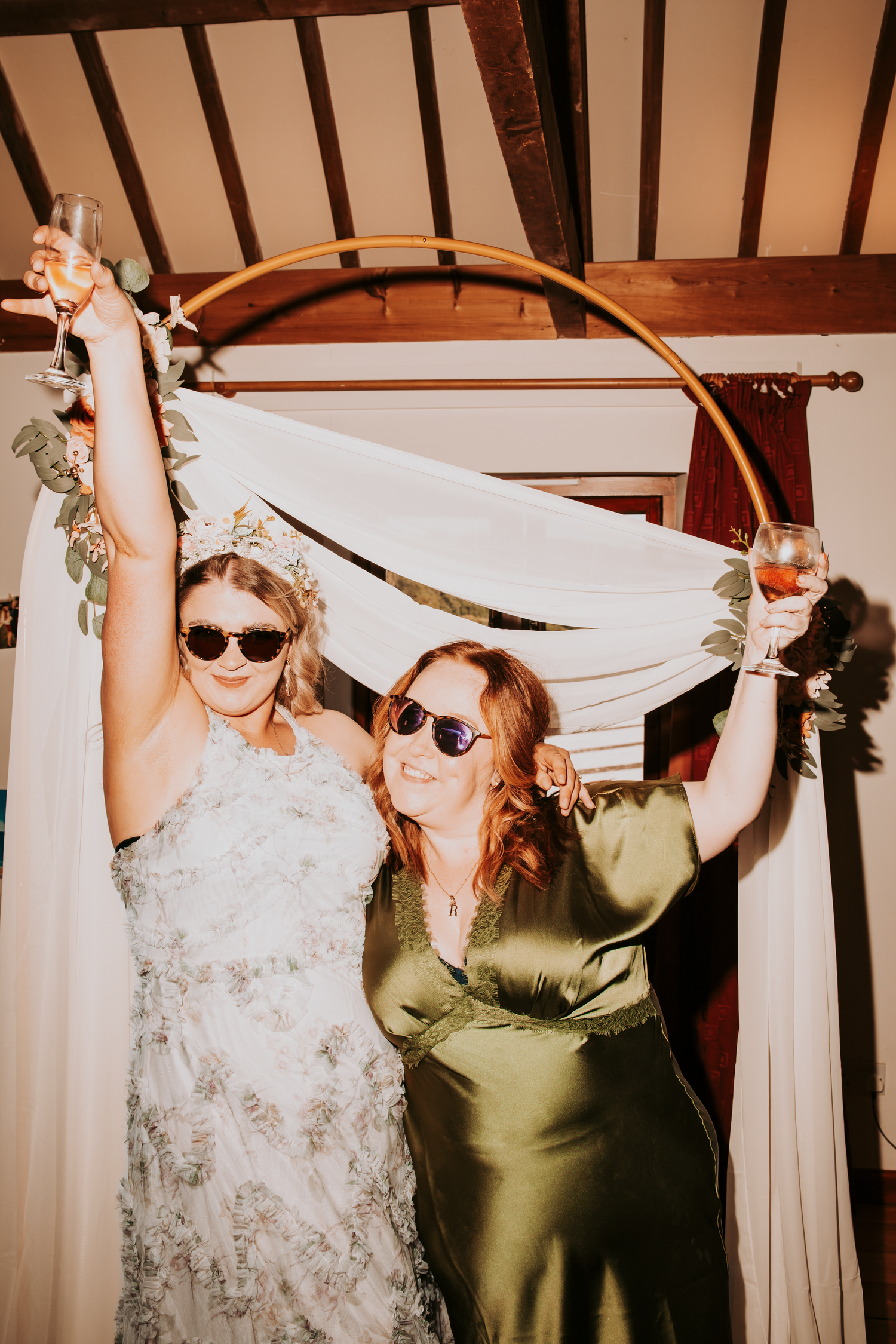 Two bridesmaids in sunglasses raise wine glasses under a golden hoop arch at a wedding reception.