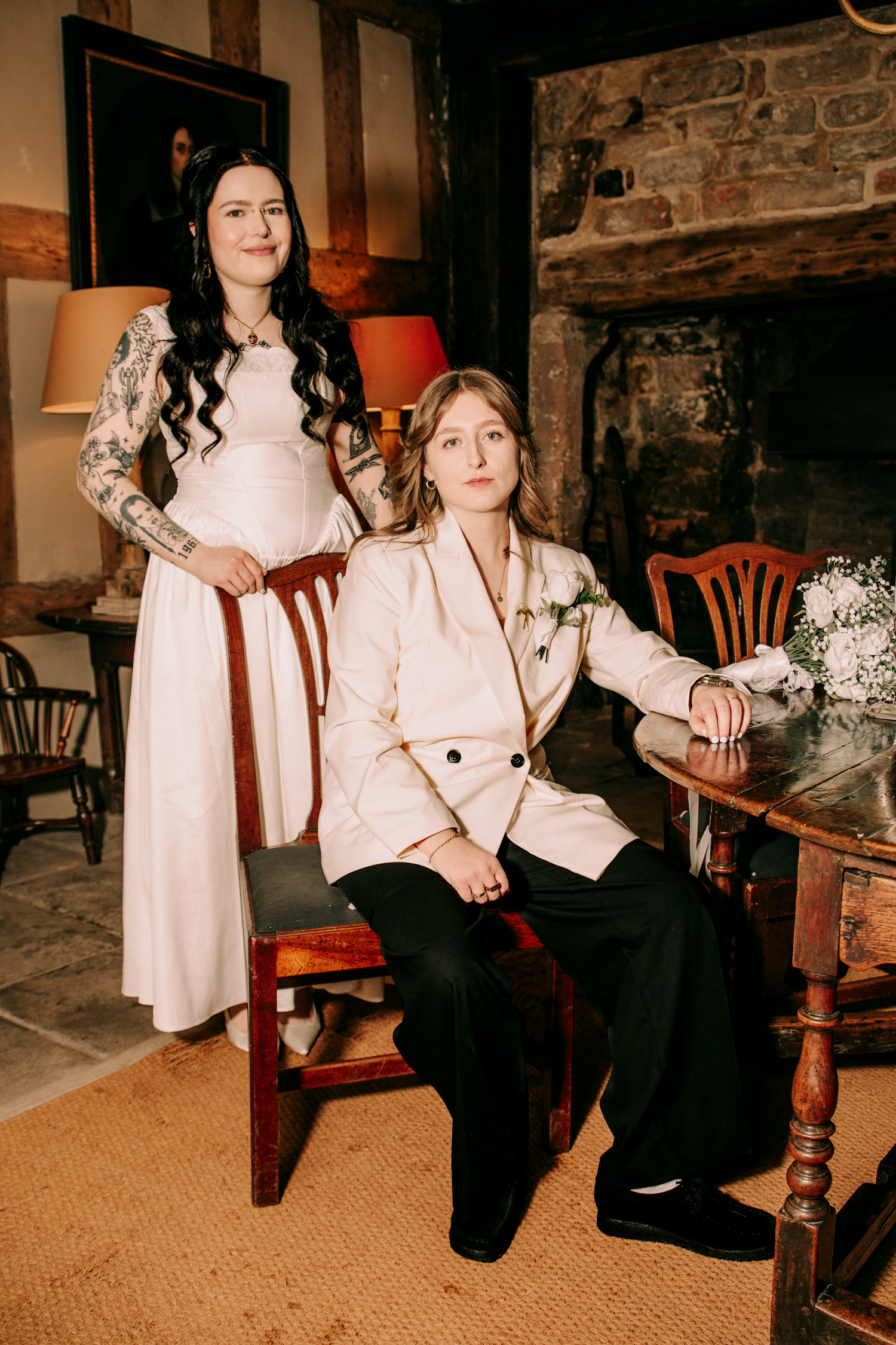Two brides pose elegantly in a rustic interior, one in a white gown and one in a cream blazer with black trousers.