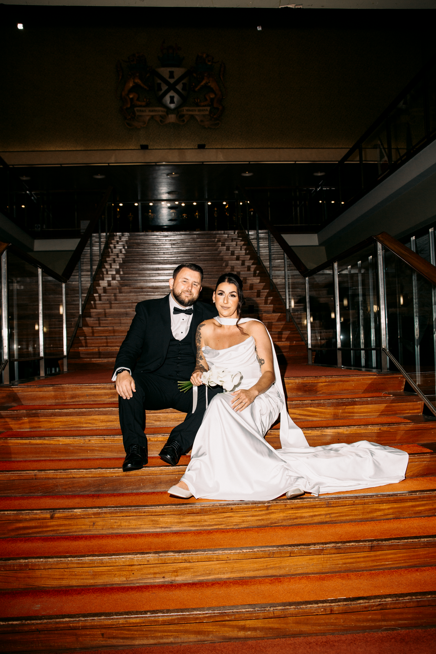 Bride and groom pose elegantly on wide wooden staircase inside grand wedding reception venue.