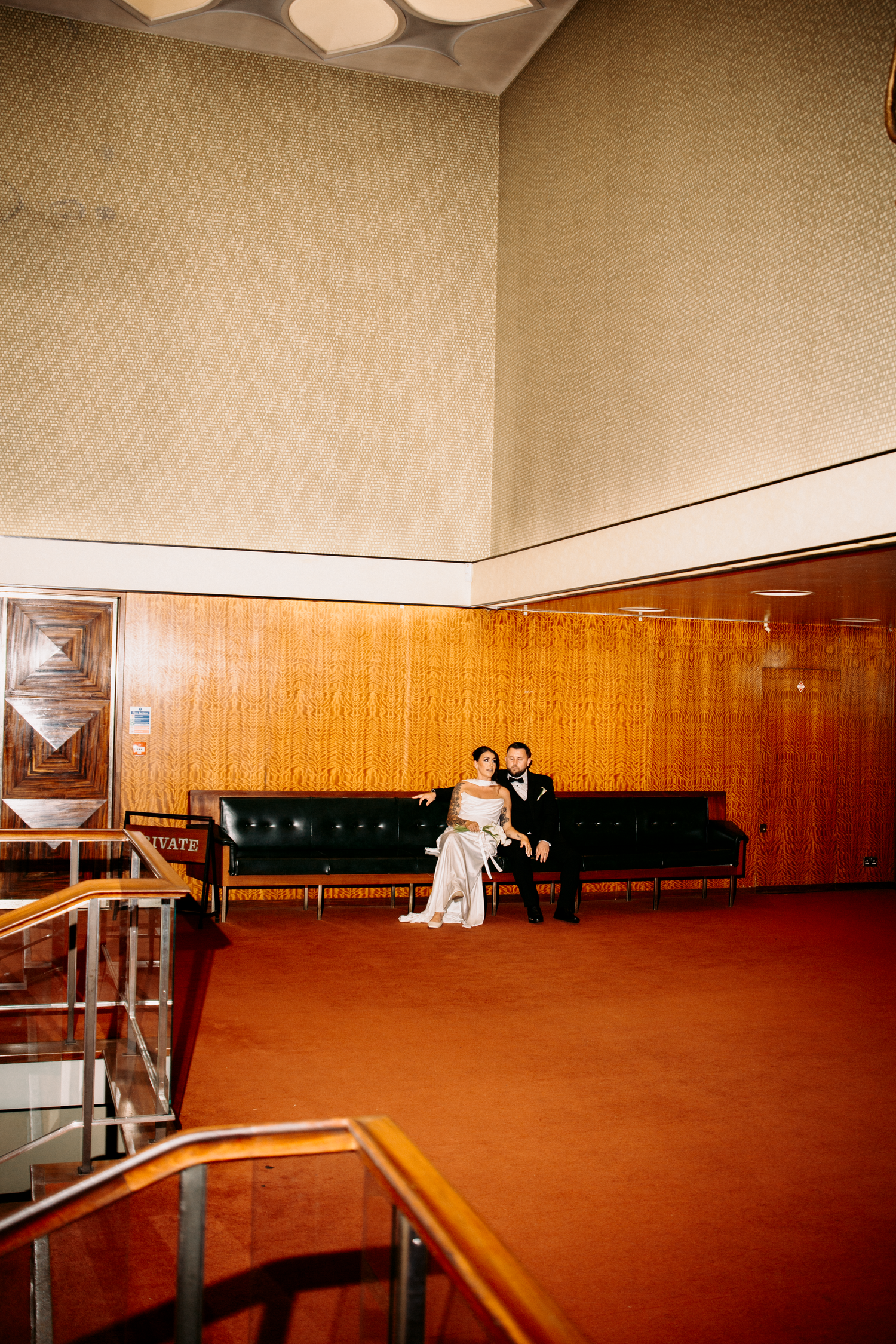 Bride and groom pose on green leather sofa in a retro-styled venue with wood-paneled walls.